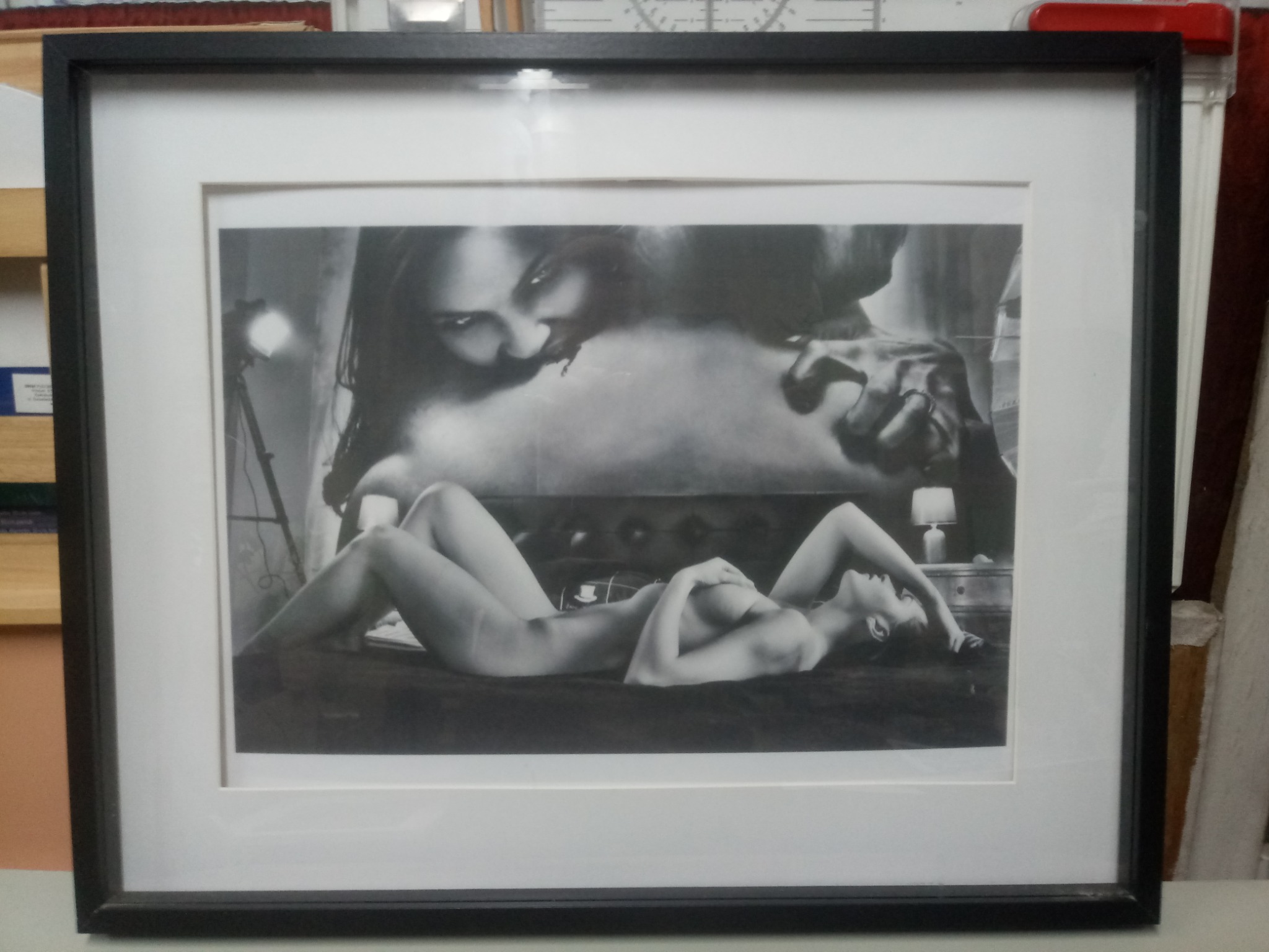 Czarno-biała fotografia artystyczna w czarnej ramie przedstawiająca leżącą nagą kobietę i twarz kobiety w tle, z widocznym oświetleniem studyjnym po lewej stronie.