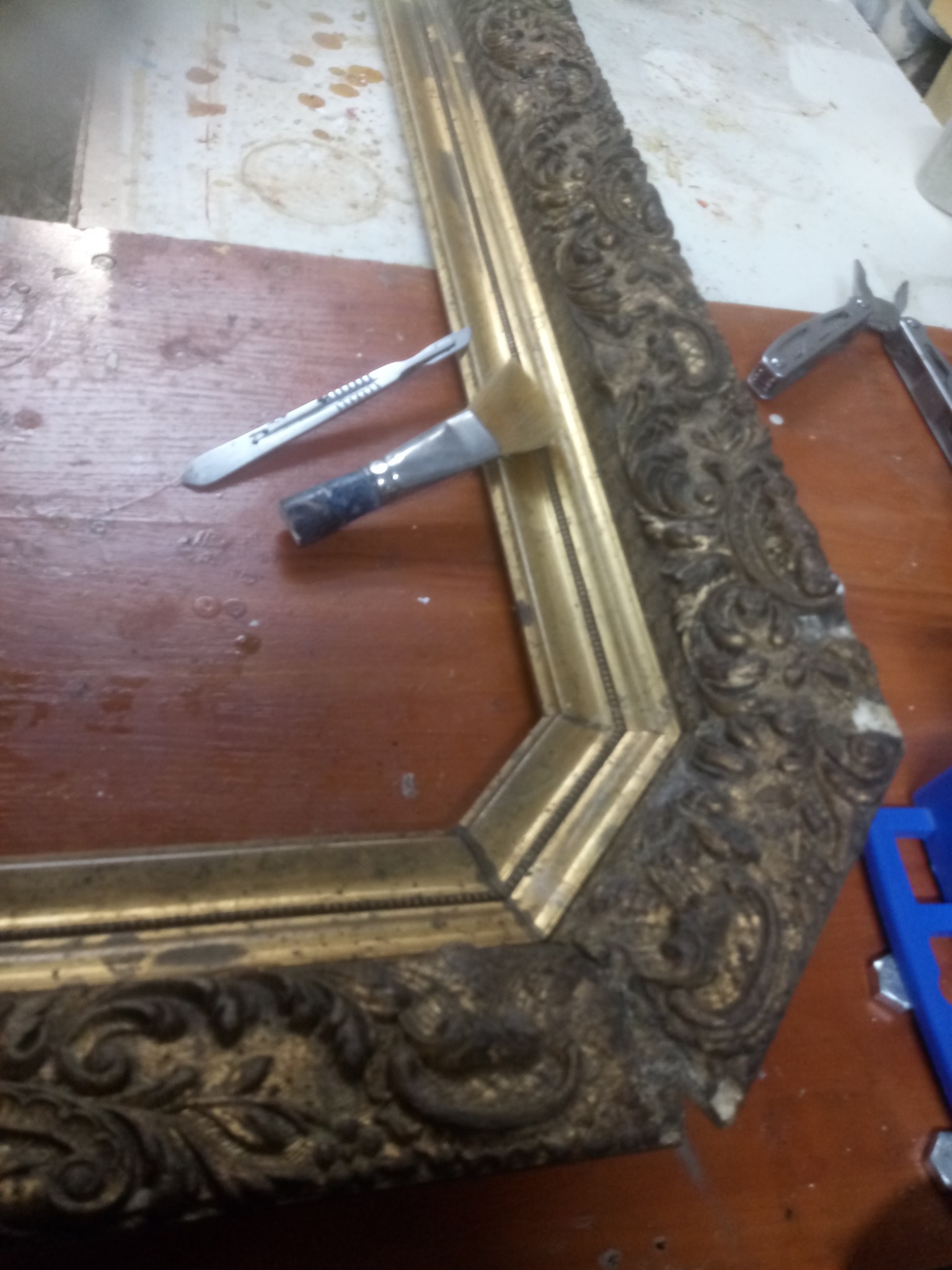 Fragment złoconej ramy z ornamentami, narzędzia konserwatorskie: pędzel i skalpel leżące na drewnianym blacie.