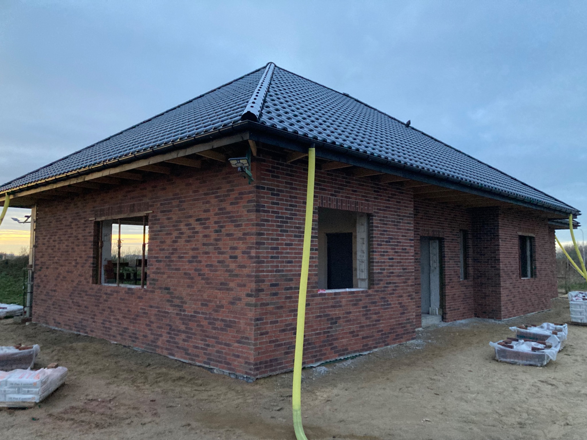 Niedokończony dom jednorodzinny z ciemnoczerwoną elewacją z cegły klinkierowej i czarnym dachem z dachówki ceramicznej, z widocznymi paczkami cegieł na ziemi i żółtym wężem na elewacji, w pochmurny...