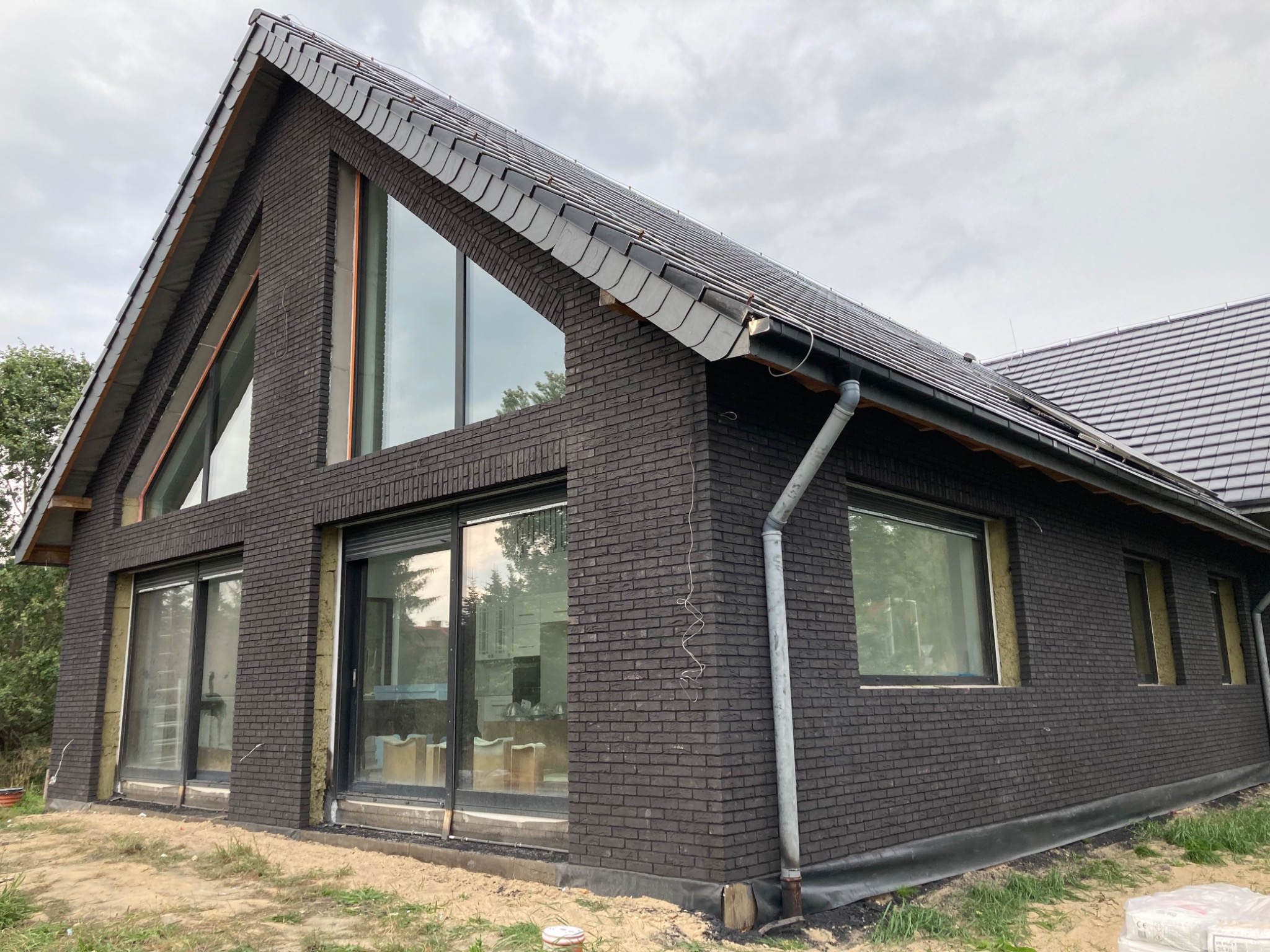 Nowoczesny dom z ciemnej cegły z dużymi oknami, widoczna izolacja wokół okien, dach pokryty ciemną dachówką, rynna odprowadzająca wodę.