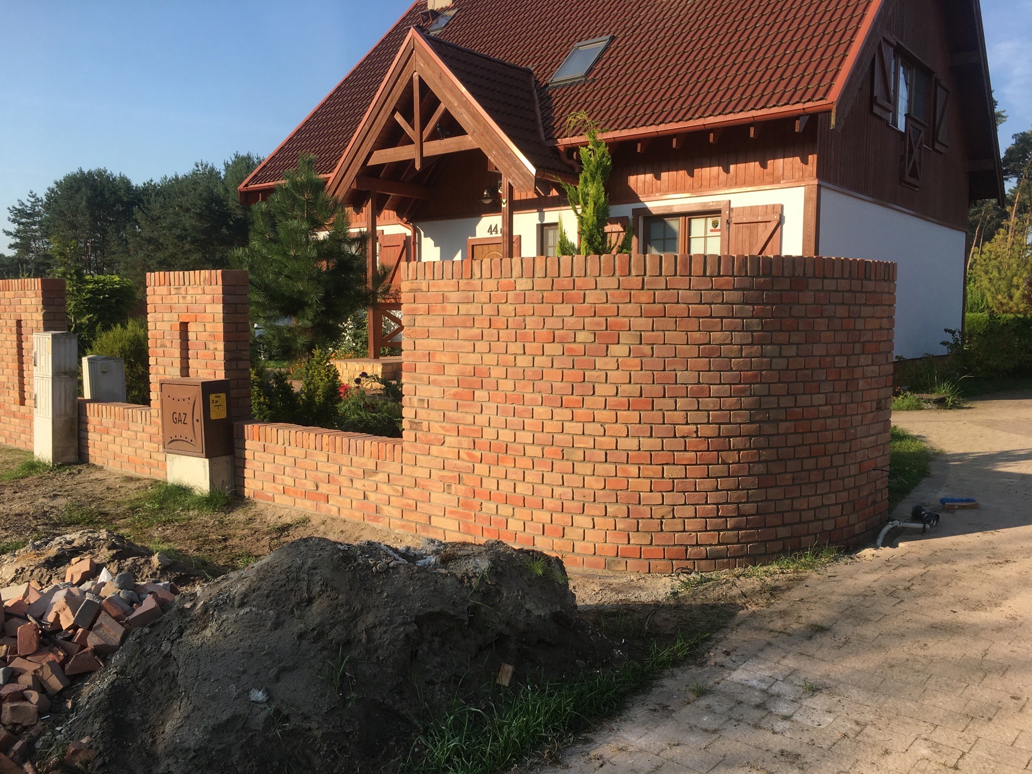 Nowo wybudowany, zaokrąglony mur z czerwonej cegły przed domem z drewnianą elewacją, widoczna sterta ziemi i porozrzucane cegły, sugerujące trwające prace wykończeniowe.