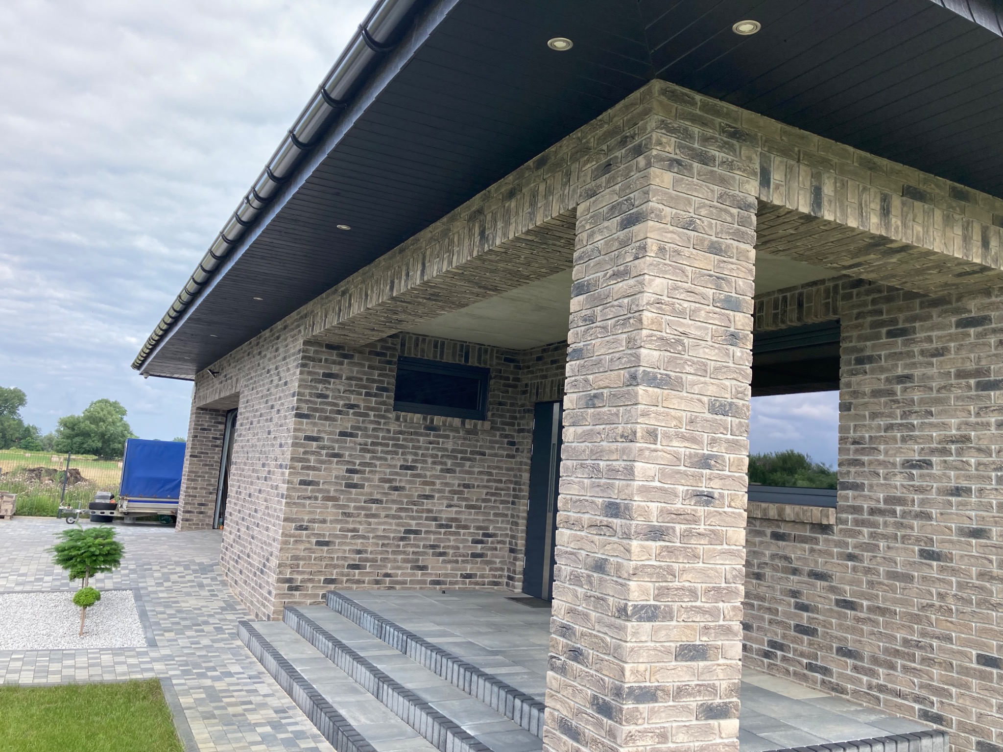 Elewacja domu jednorodzinnego z cegły klinkierowej w odcieniach szarości i beżu, widok z zewnątrz na wejście z kolumną i schodami, z ciemnym podbitką dachową oraz kostką brukową na podjeździe.