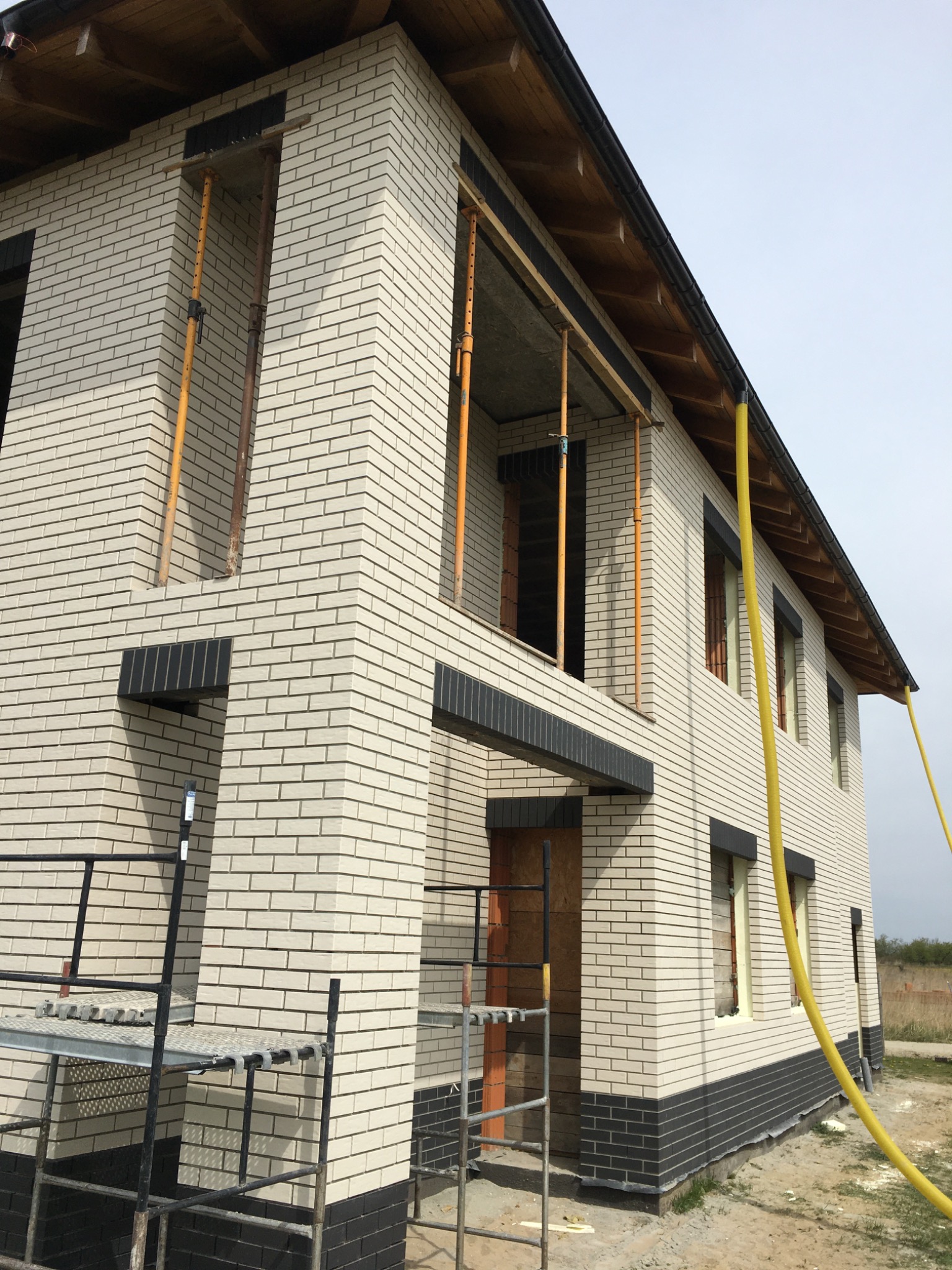 Dwukondygnacyjny budynek w trakcie murowania elewacji z jasnej cegły klinkierowej z ciemnymi akcentami, widoczne rusztowania i żółty wąż.