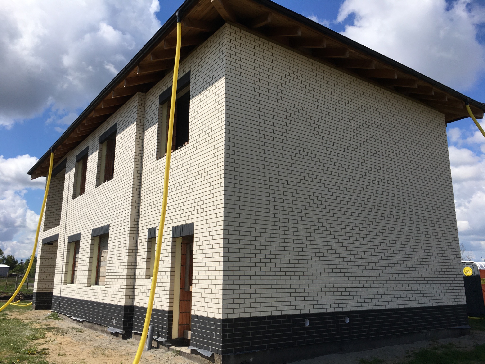 Dwupiętrowy dom w trakcie budowy, z jasną ceglaną elewacją i ciemną podmurówką, widoczne rury wentylacyjne na tle błękitnego nieba z chmurami.