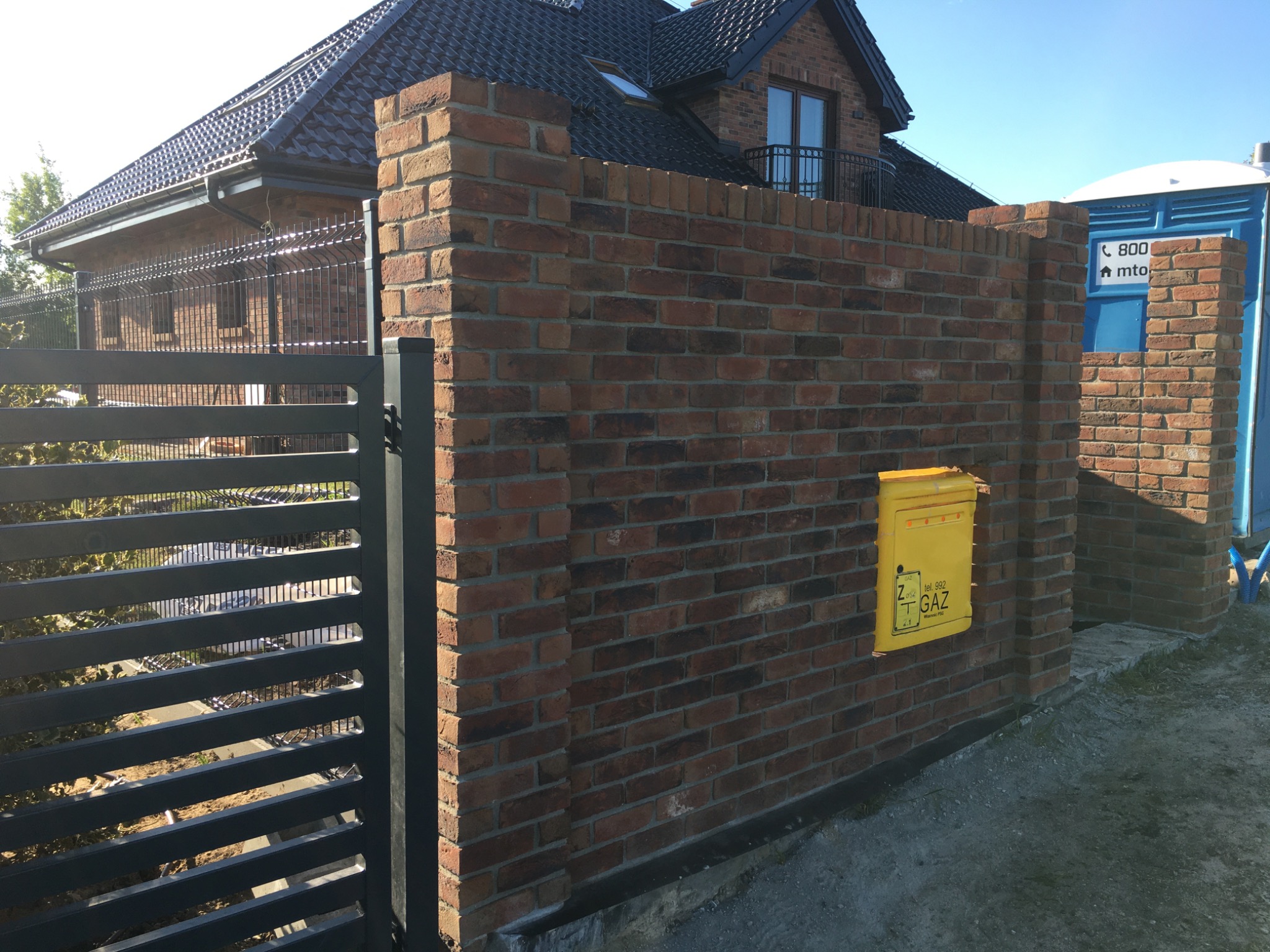Ceglany mur ogrodzeniowy z żółtą skrzynką gazową, przylegający do nowoczesnego domu z ciemną dachówką i czarnym ogrodzeniem panelowym.