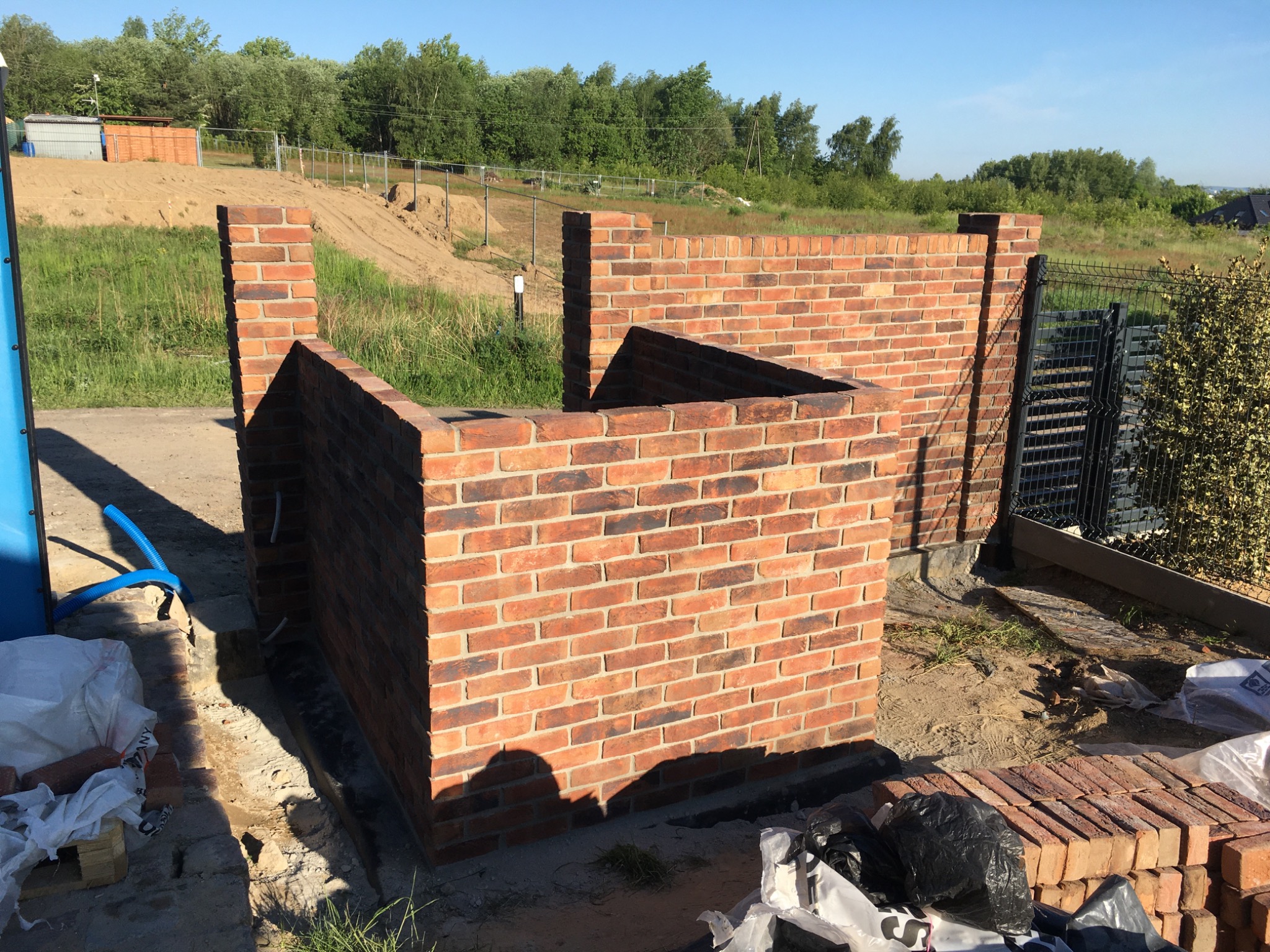 Wznoszenie muru z czerwonej cegły, widoczne fragmenty instalacji i materiały budowlane, w tle zieleń i fragmenty zabudowy.
