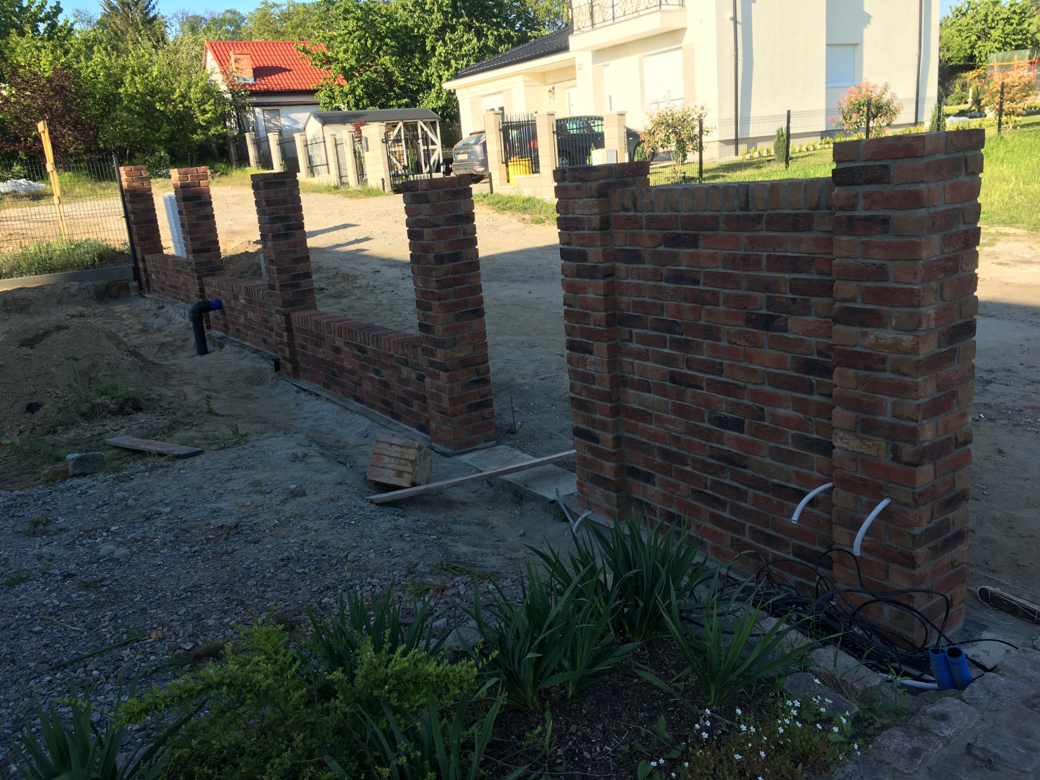 Budowa ogrodzenia z cegły klinkierowej w Stargardzie, widoczne słupki i fragment muru z wystającymi rurami instalacyjnymi, w tle dom jednorodzinny.