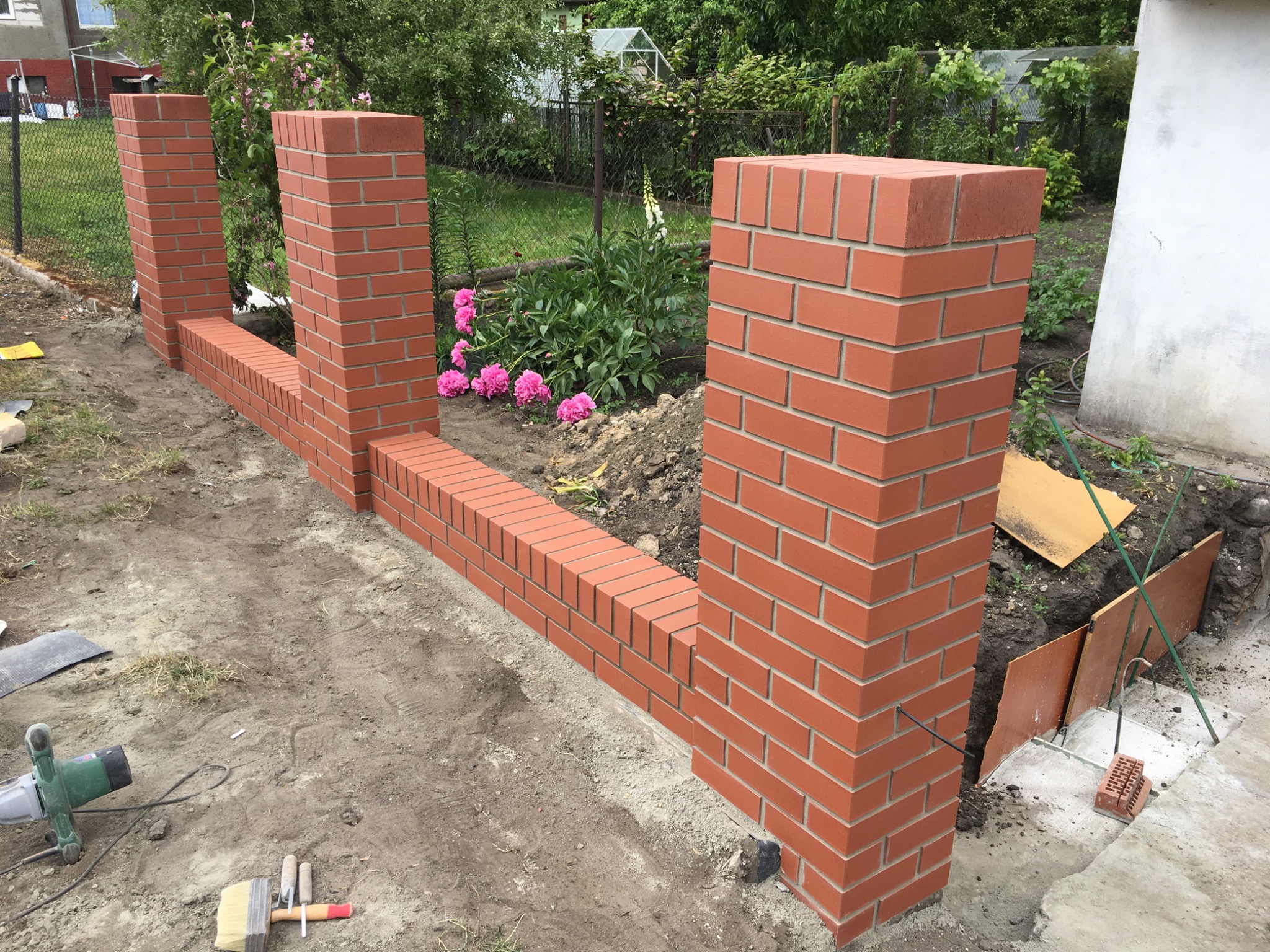 Wznoszenie murowanego ogrodzenia z czerwonej cegły z widocznymi narzędziami murarskimi i kwiatami w tle.