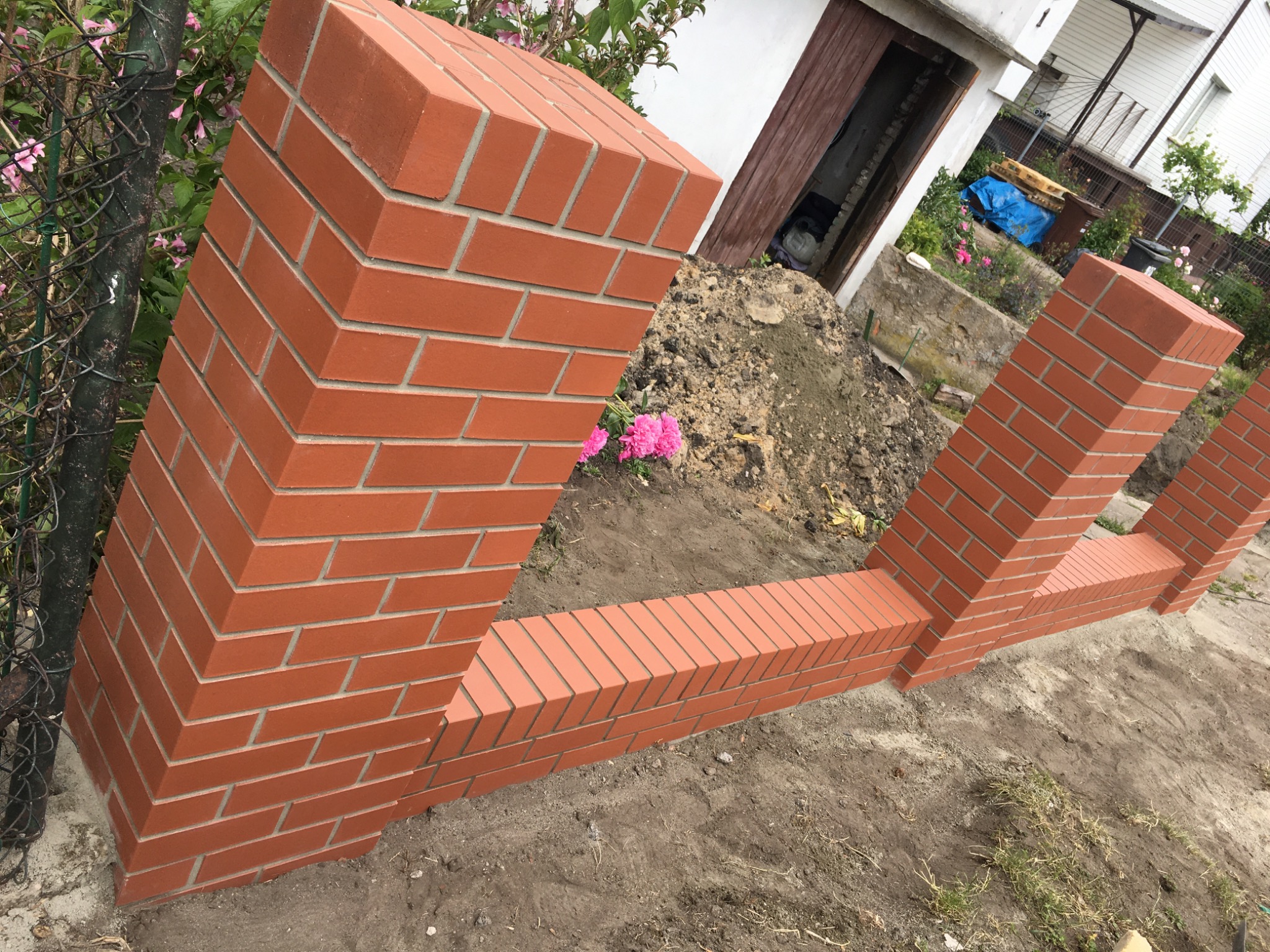 Świeżo wymurowane, ceglane słupki ogrodzeniowe z elementami podmurówki, widoczne na tle ogrodu z kwiatami i fragmentem budynku.