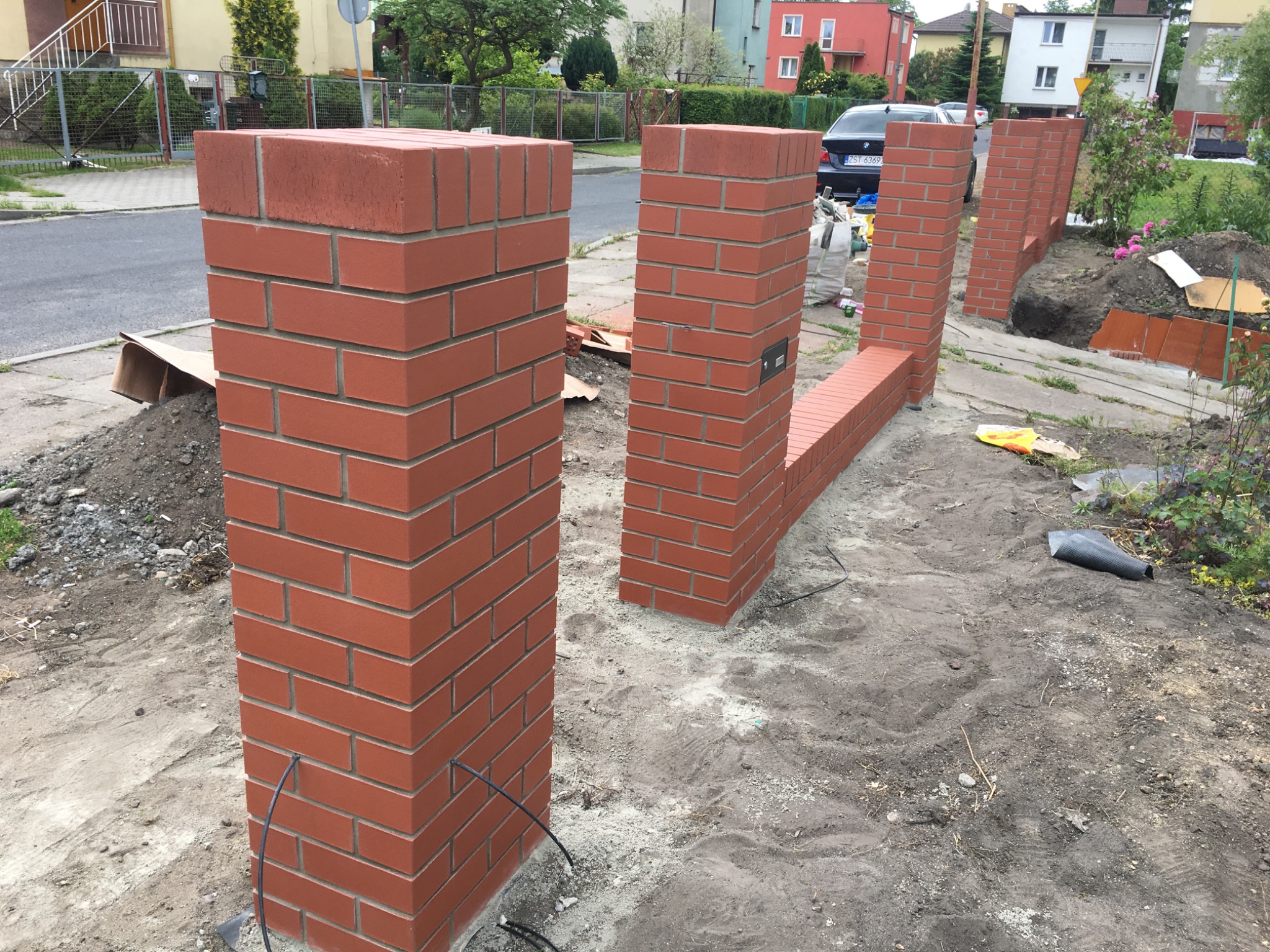 Wznoszenie ogrodzenia z czerwonej cegły klinkierowej, widoczne słupki i fragment muru w trakcie budowy, przewody elektryczne wystające ze słupka, otoczenie w stanie surowym.