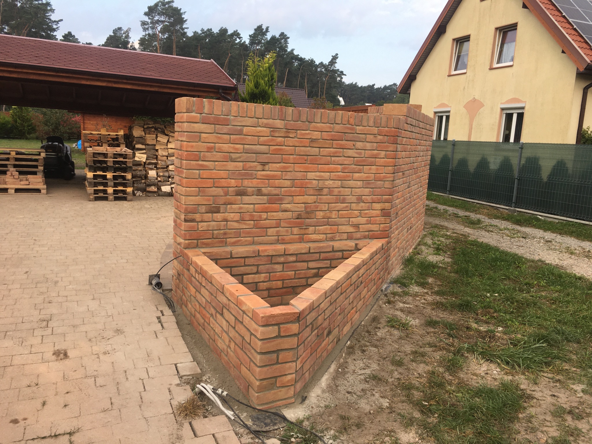 Wznoszona z cegły klinkierowej konstrukcja w kształcie litery U, prawdopodobnie fragment ogrodzenia lub element małej architektury ogrodowej, widoczny na tle domu i zadaszenia z drewnem.