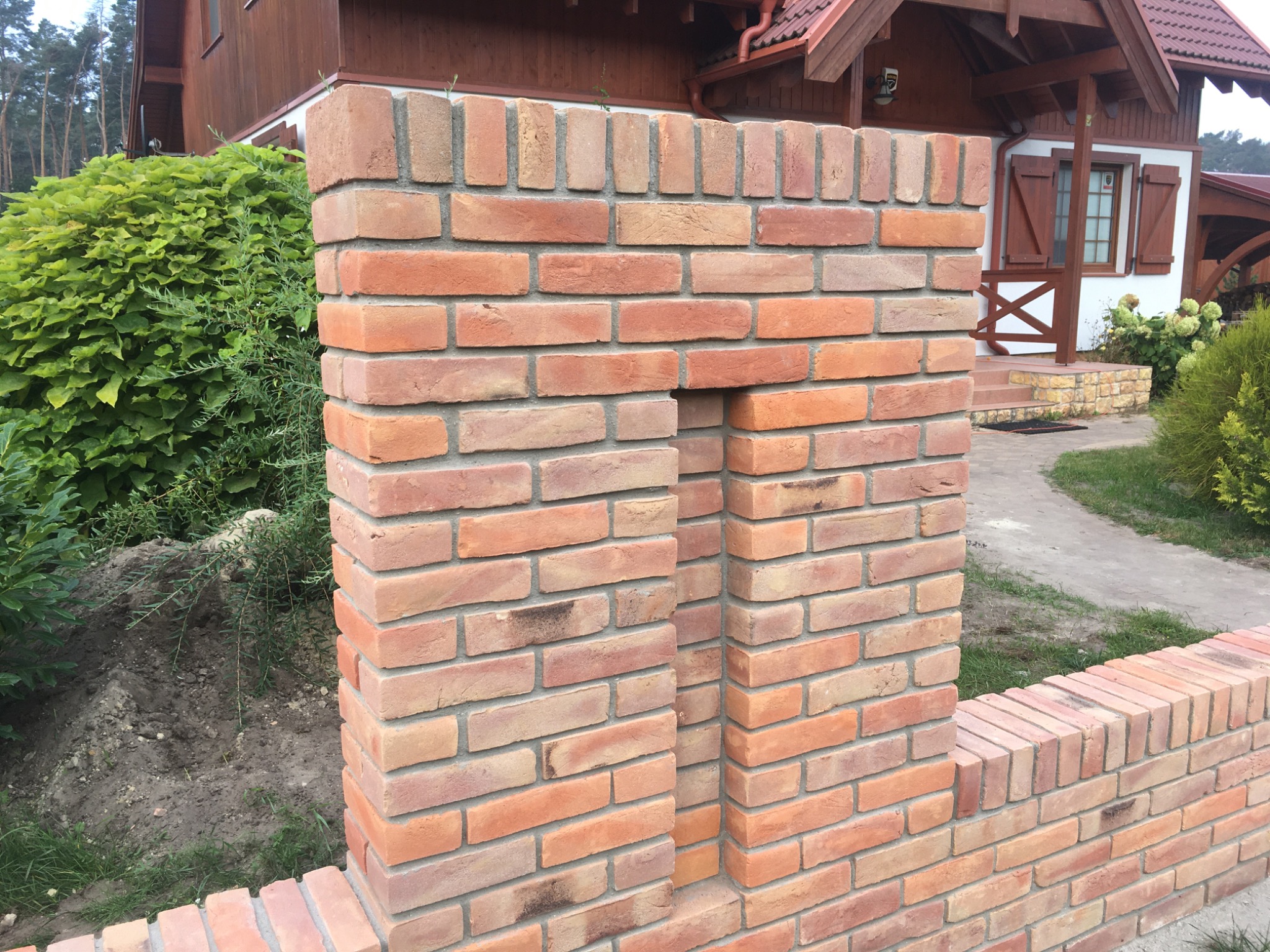 Fragment ogrodzenia z cegły klinkierowej z charakterystycznym otworem, na tle domu z drewnianymi elementami i zielenią.