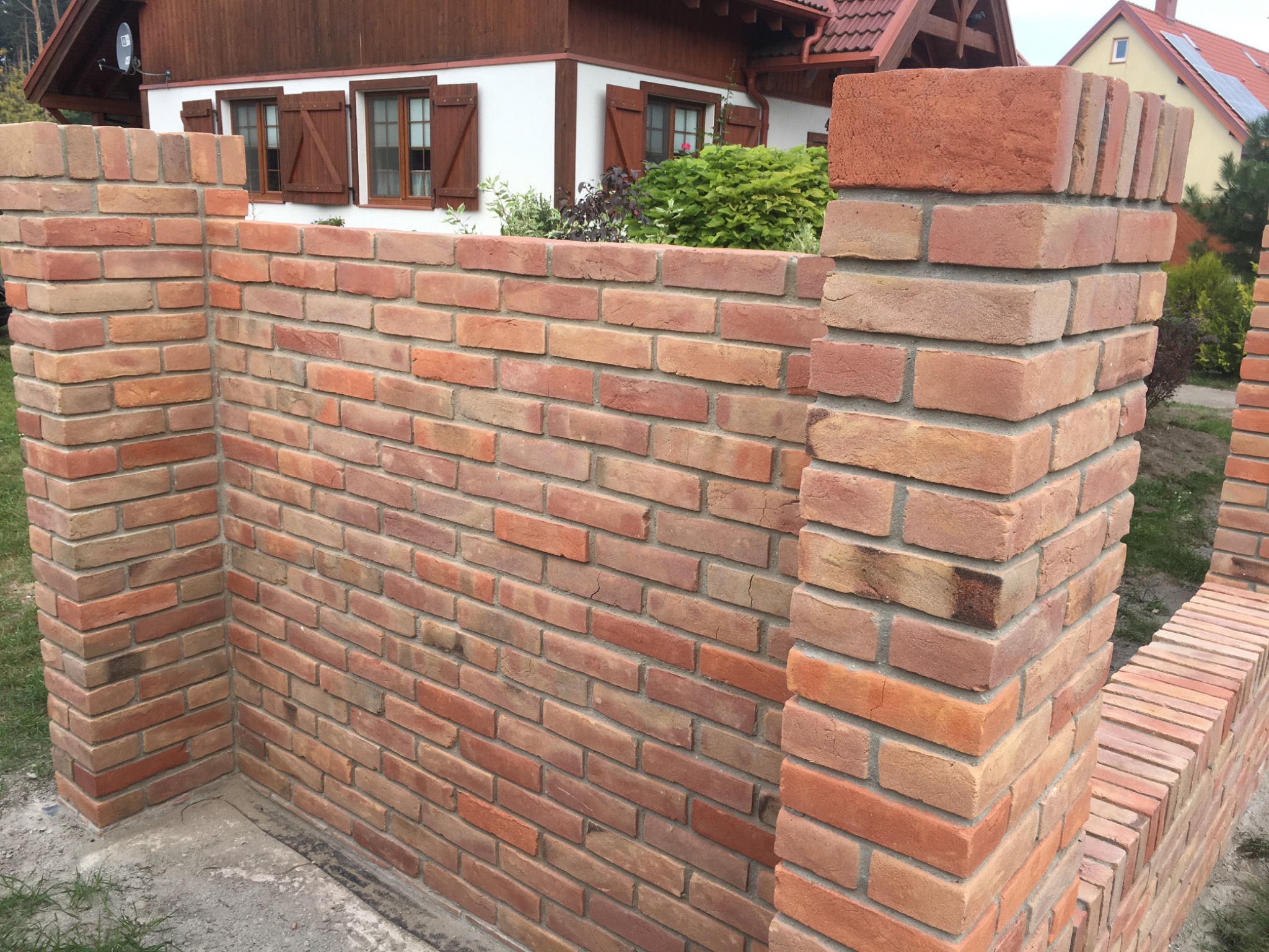 Nowo wybudowany, ceglany murek ogrodzeniowy z czerwonej cegły klinkierowej, z widoczną fakturą i spoinami, na tle domu z drewnianymi okiennicami.