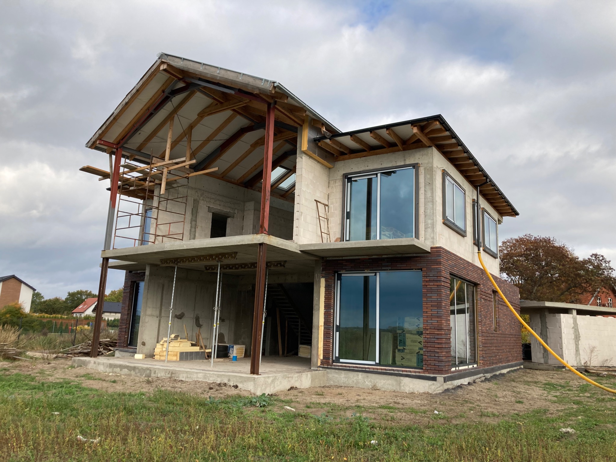 Budynek w stanie surowym otwartym z częściowo wykonaną elewacją z cegły klinkierowej, widoczne rusztowania i elementy konstrukcyjne dachu.