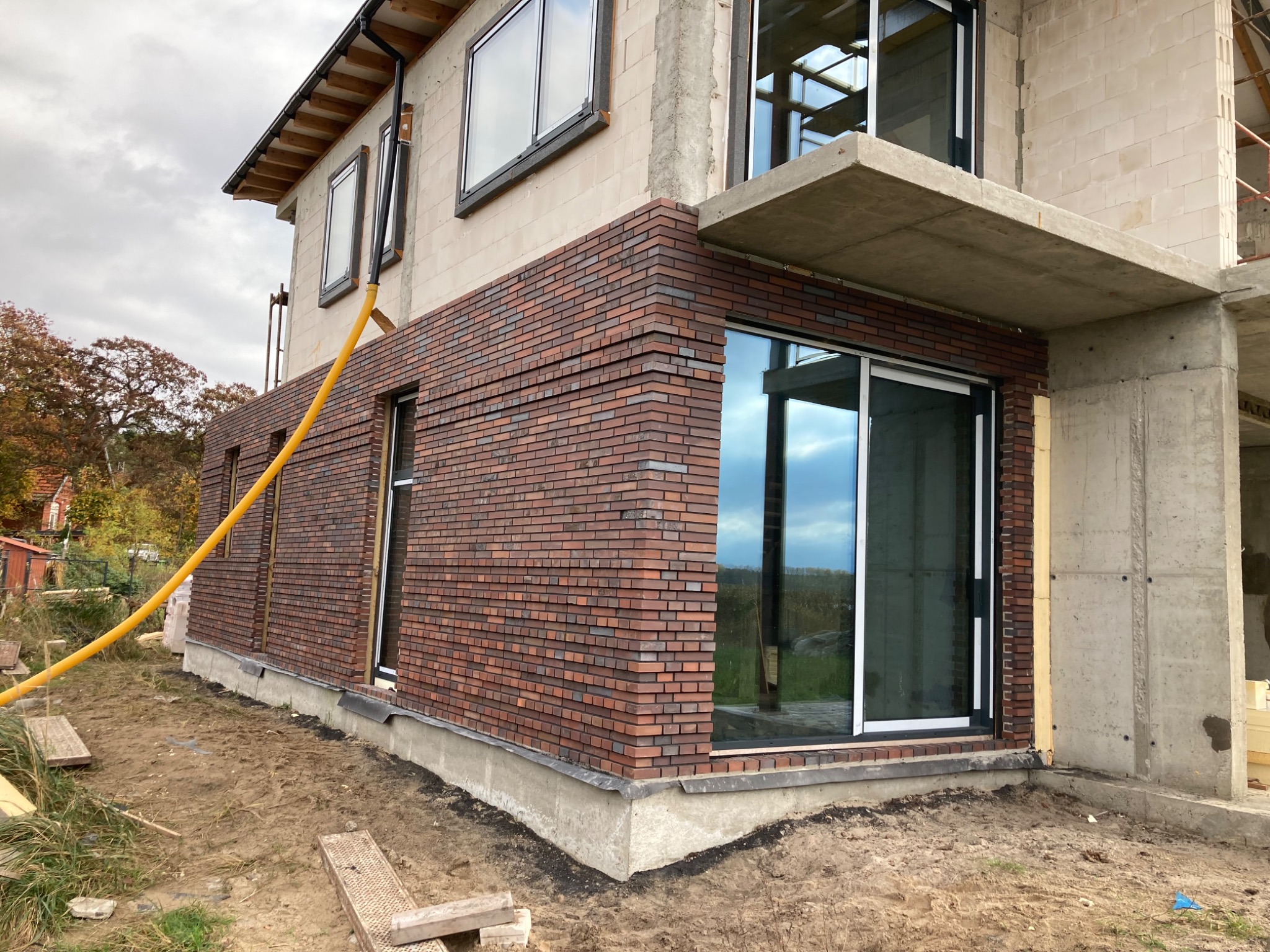 Budynek w trakcie wykańczania z elewacją z cegły klinkierowej; widoczny żółty wąż na elewacji; surowy beton na balkonie; okna z czarnymi ramami.