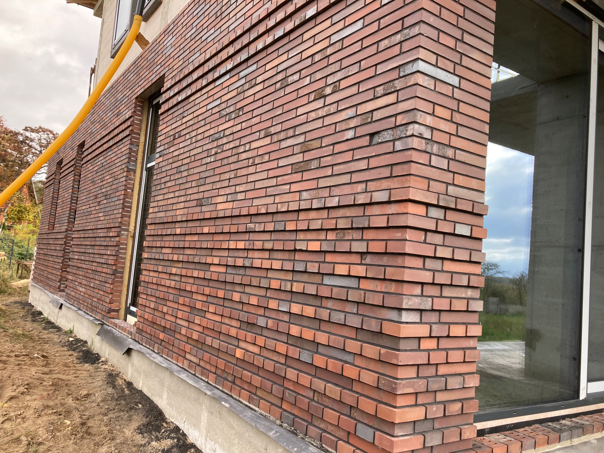 Fragment elewacji budynku z cegły klinkierowej w odcieniach czerwieni i brązu, z widocznym oknem i żółtą rurą spustową, ukazujący detal wykończenia i fakturę materiału.