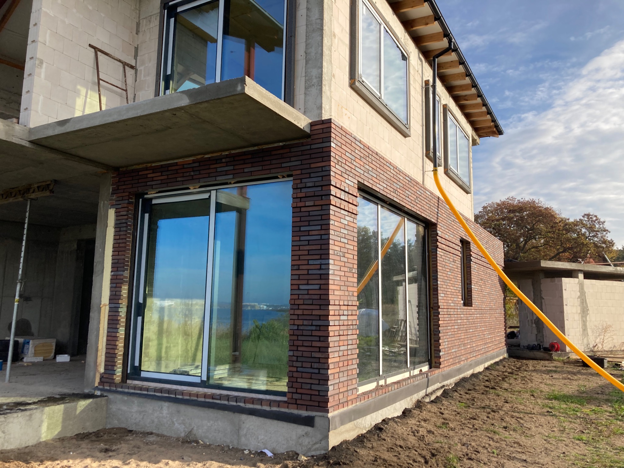 Budynek w trakcie wykańczania elewacji z cegły klinkierowej, widoczne duże okna z odbiciem krajobrazu, żółty wąż na elewacji, budynek w stanie surowym otwartym.