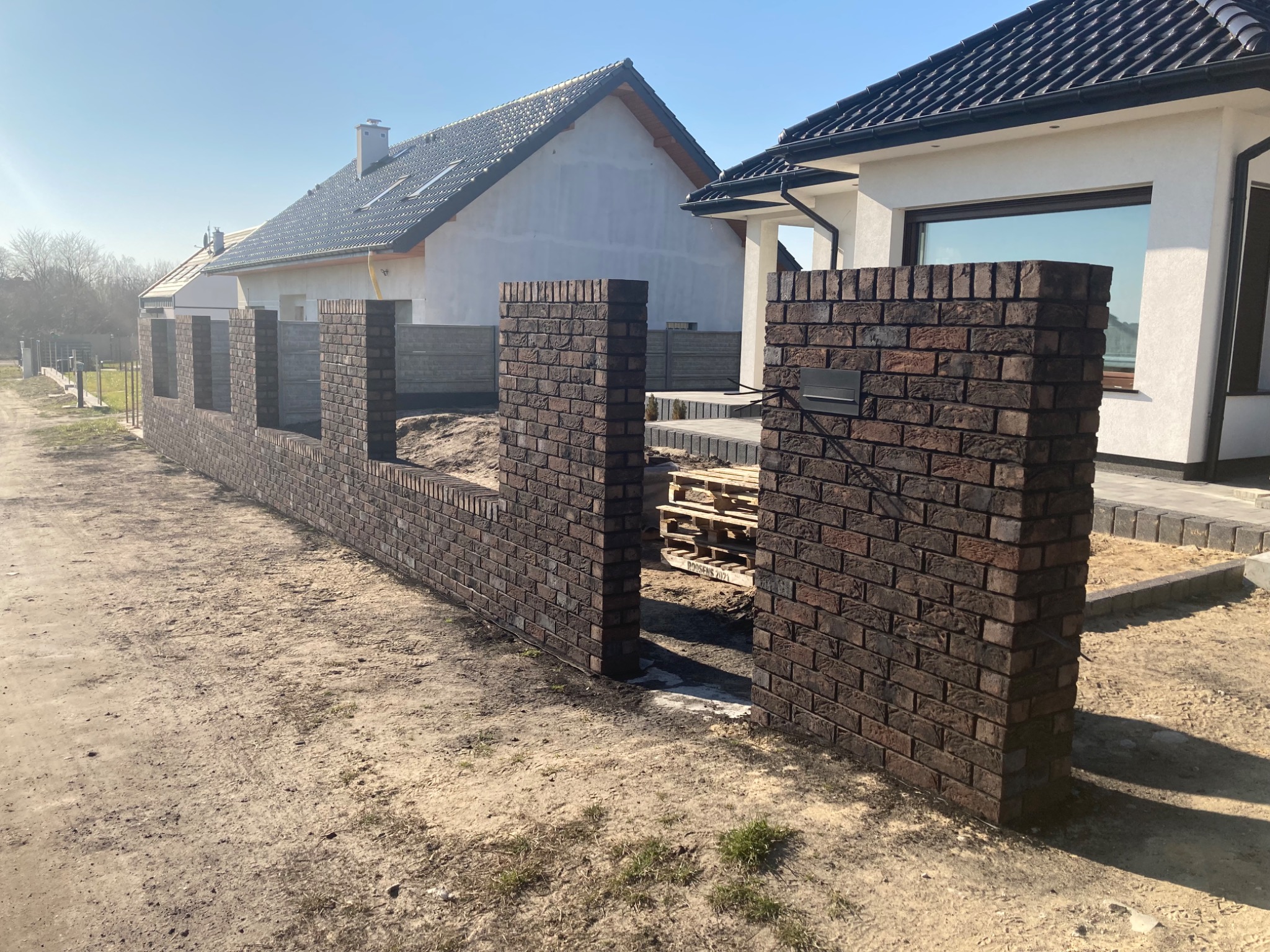 Nowoczesne ogrodzenie z ciemnej cegły klinkierowej w trakcie budowy, widoczne palety z cegłami i fragmenty domów w tle.