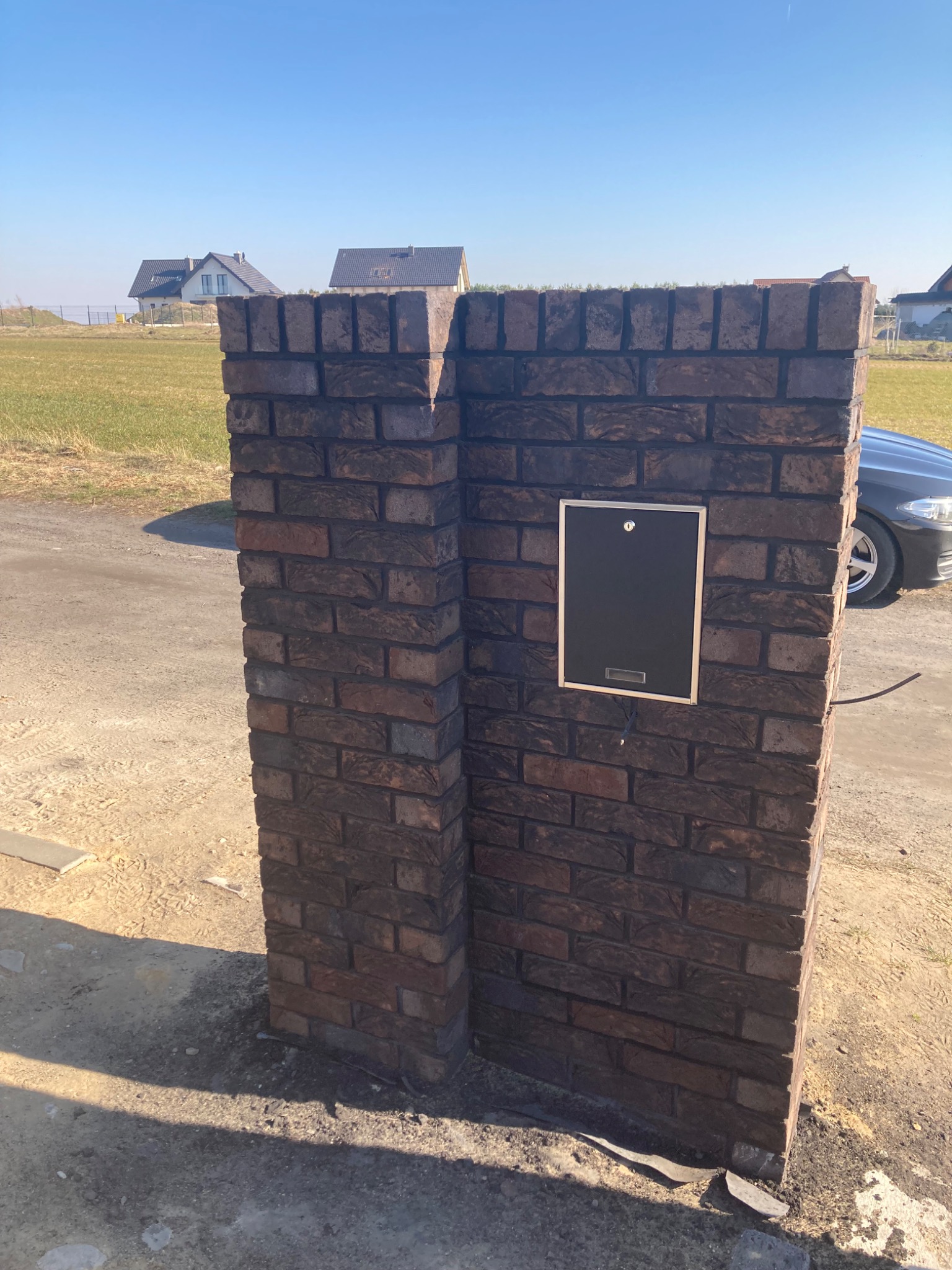 Murowany słupek z cegły klinkierowej z zamontowaną czarną skrzynką pocztową, widoczne domy w oddali i fragment samochodu.