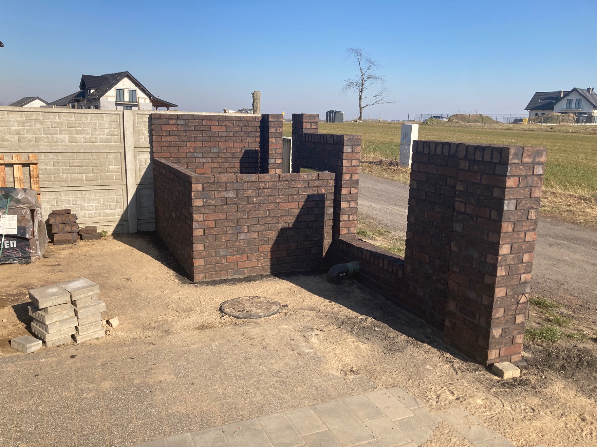 Wznoszenie muru z cegły klinkierowej na posesji, widoczne fragmenty konstrukcji, stos cegieł i elementów betonowych, w tle domy jednorodzinne i błękitne niebo.