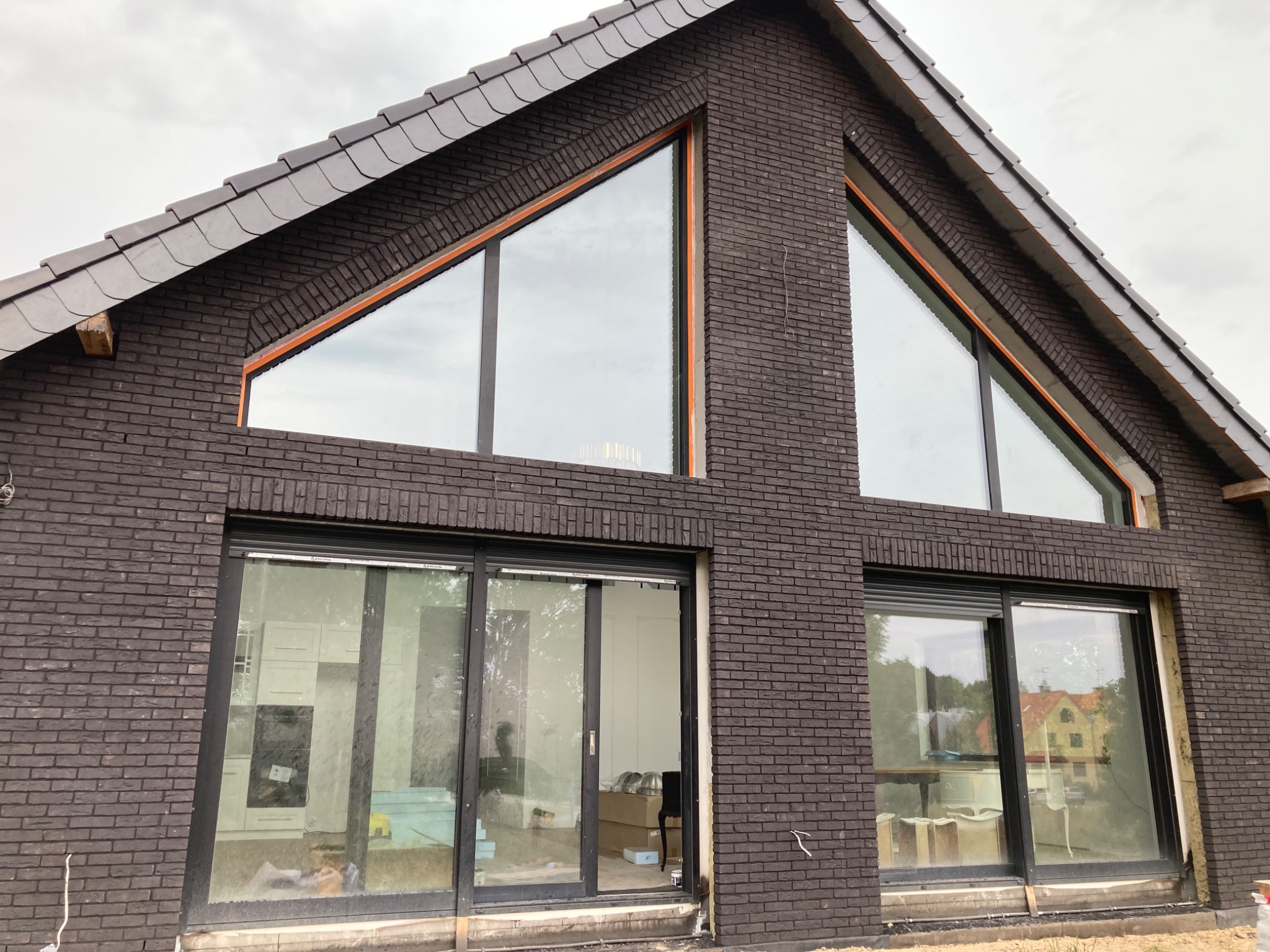 Nowoczesny dom z ciemnej cegły z dużymi, trójkątnymi oknami i przesuwnymi drzwiami balkonowymi, widoczne prace wykończeniowe wokół okien.