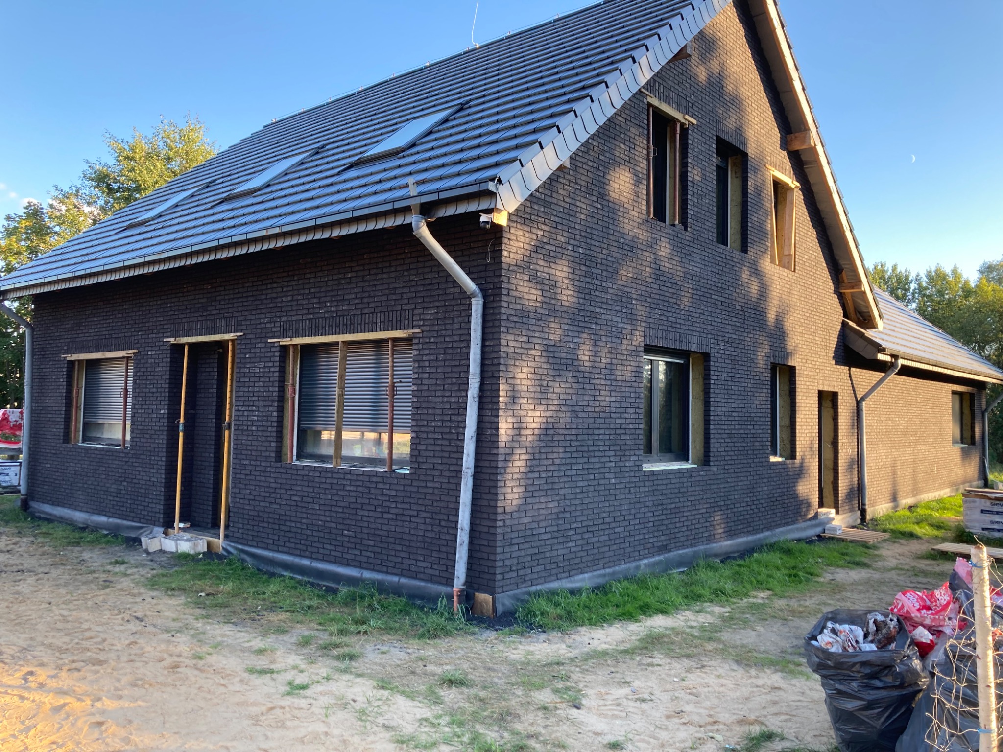 Dom jednorodzinny w trakcie budowy, elewacja z ciemnej cegły klinkierowej, widoczne okna z żaluzjami i otwory okienne bez ram, dach pokryty szarą dachówką, teren wokół domu częściowo...