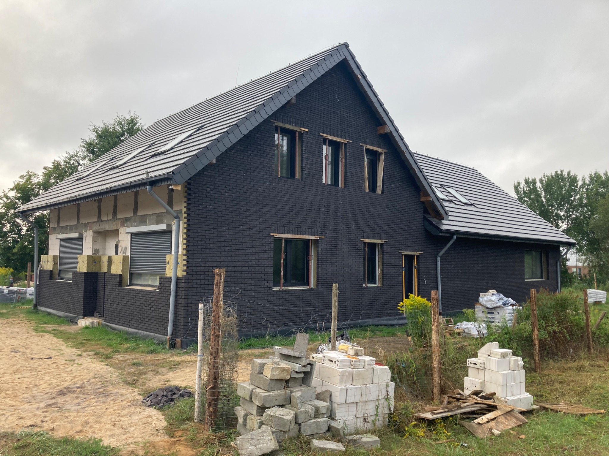 Dom w trakcie budowy z czarną elewacją z cegły klinkierowej i szarą dachówką, widoczne materiały budowlane i fragmenty ocieplenia ścian.