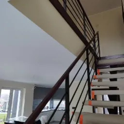 Metalowa balustrada schodowa z brązowymi elementami, widok z dołu, schody zabezpieczone folią malarską, w tle jasna kuchnia i okno.