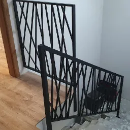 Nowoczesna, czarna balustrada schodowa z geometrycznym wzorem w pionowe, przecinające się linie, widoczna z góry, z podłogą imitującą drewno w tle.