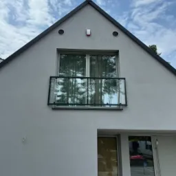 Nowoczesny dom z balkonem z balustradą ze szkła hartowanego, odbijającą drzewa i niebo, widoczne detale architektoniczne jak wentylacja i czujnik alarmowy.