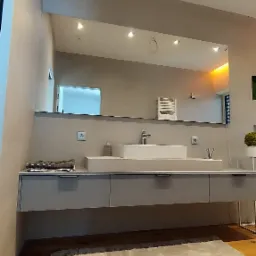 Nowoczesna łazienka z dużym lustrem nad umywalką wpuszczoną w szarą szafkę, jasna podłoga i puszysty dywanik.