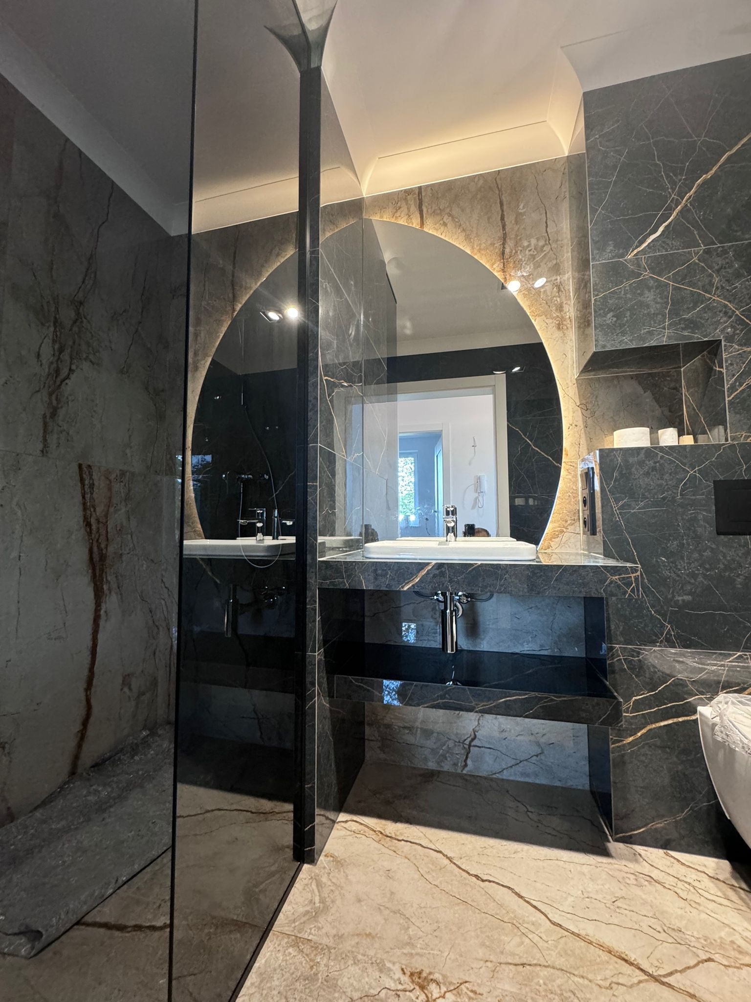 Nowoczesna łazienka z marmurowymi ścianami i podświetlanym, okrągłym lustrem nad umywalką, oddzielona szklaną ścianką prysznicową.