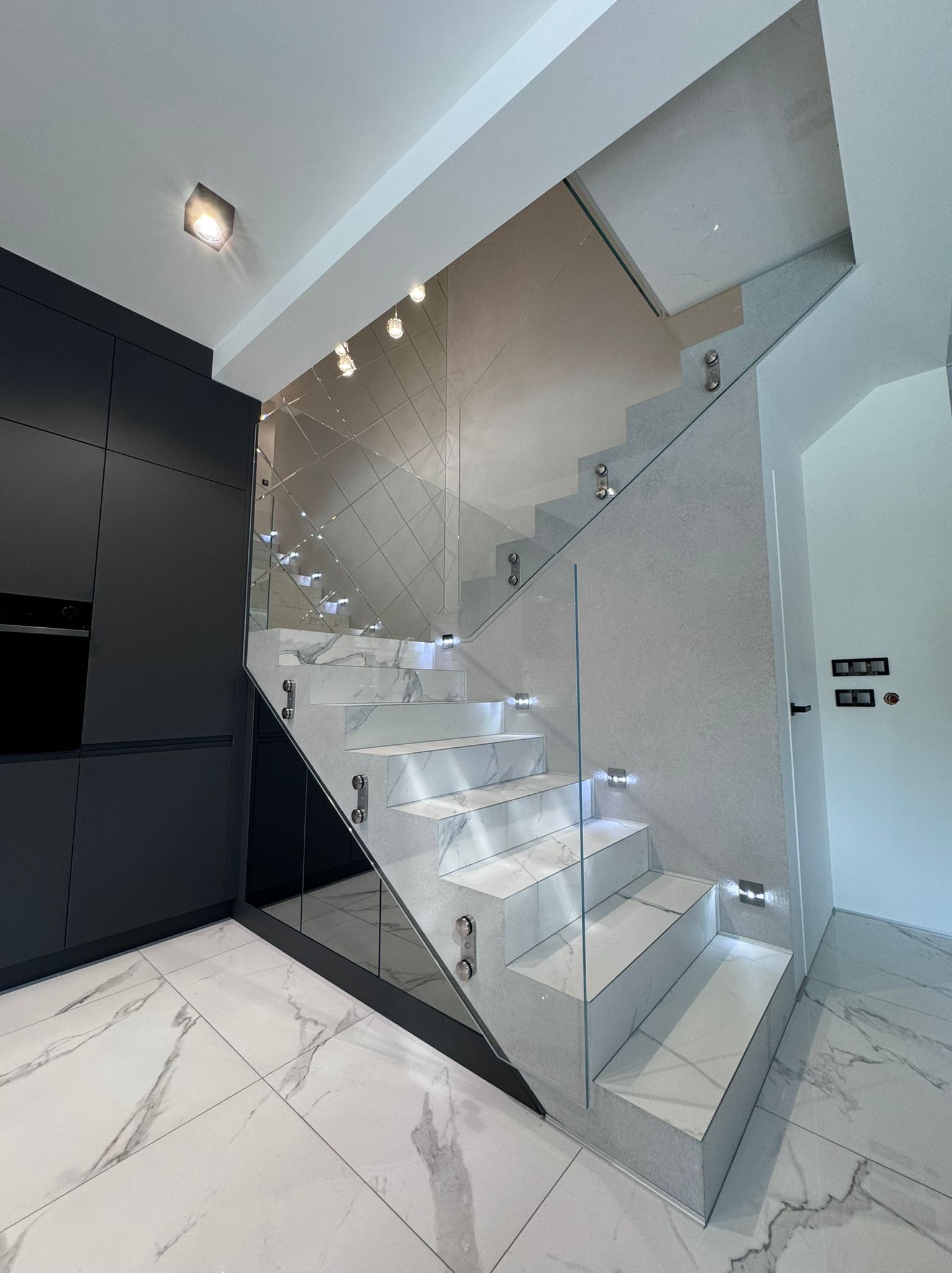 Nowoczesne schody z marmurowymi stopniami i szklaną balustradą, oświetlone punktowymi lampkami LED, przylegające do czarnej zabudowy meblowej w minimalistycznym wnętrzu.