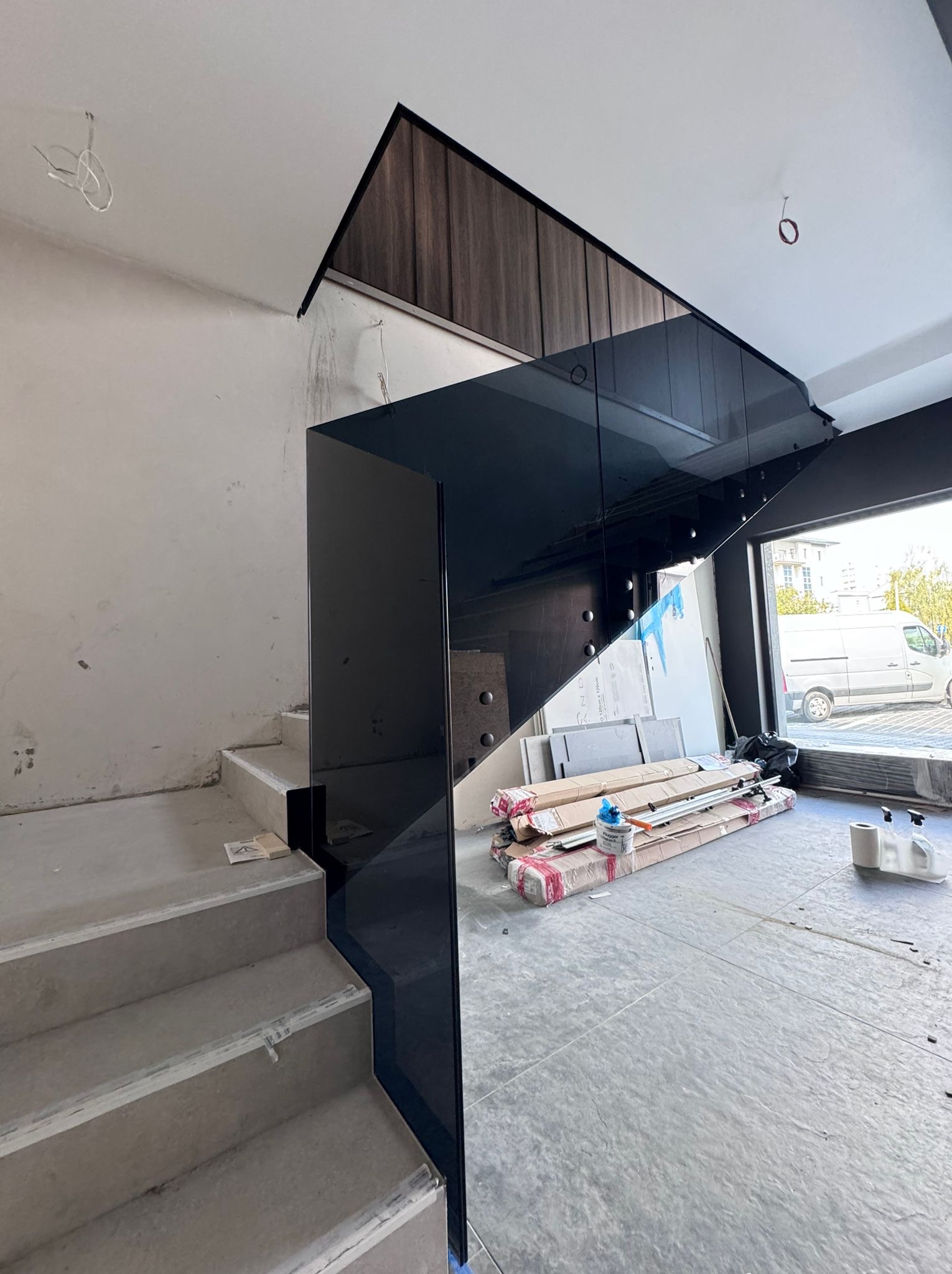 Montaż szklanej balustrady schodowej w surowym wnętrzu, widoczne materiały budowlane i elementy montażowe na podłodze.