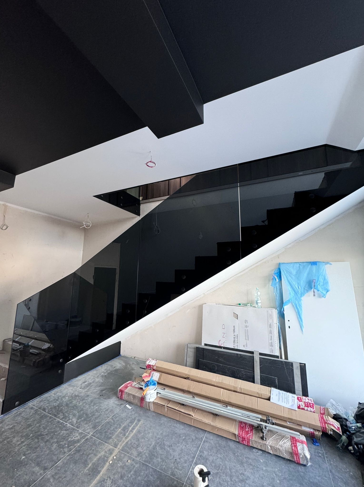 Montaż czarnej, szklanej balustrady schodowej w nowoczesnym wnętrzu z czarnym sufitem; widoczne materiały budowlane i narzędzia na podłodze.