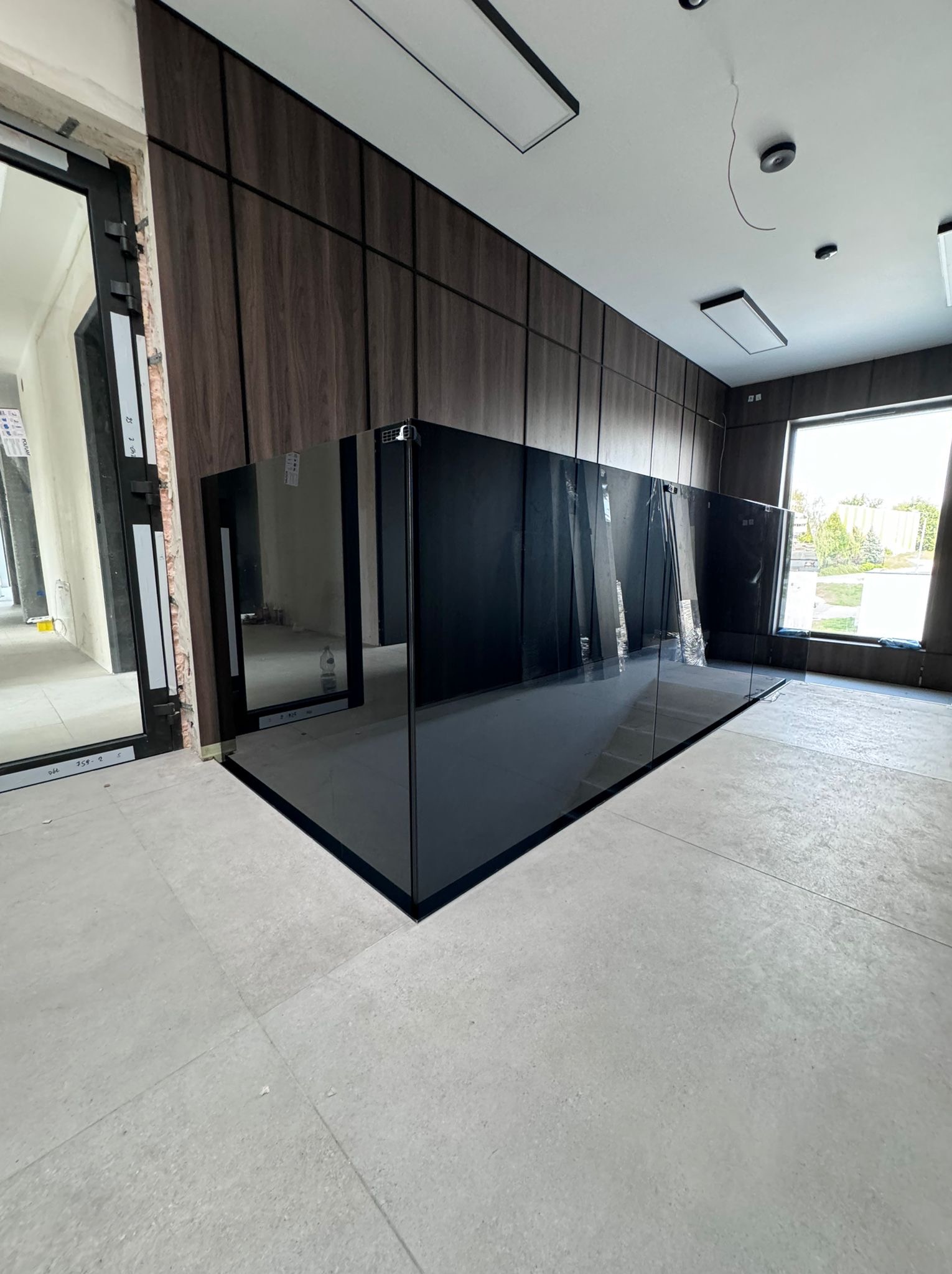 Ciemna, szklana balustrada w nowoczesnym wnętrzu z panelami ściennymi z imitacji drewna, widoczna przez okno zieleń, surowy stan wykończenia podłogi.