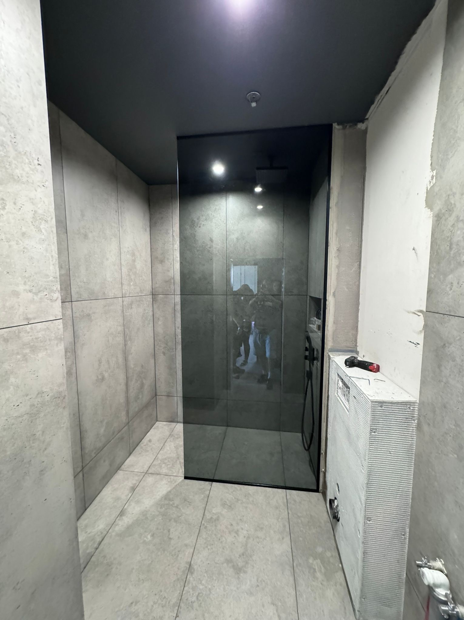 Montaż szklanej kabiny prysznicowej w łazience wykończonej płytkami imitującymi beton, widoczny poziomica i narzędzia.