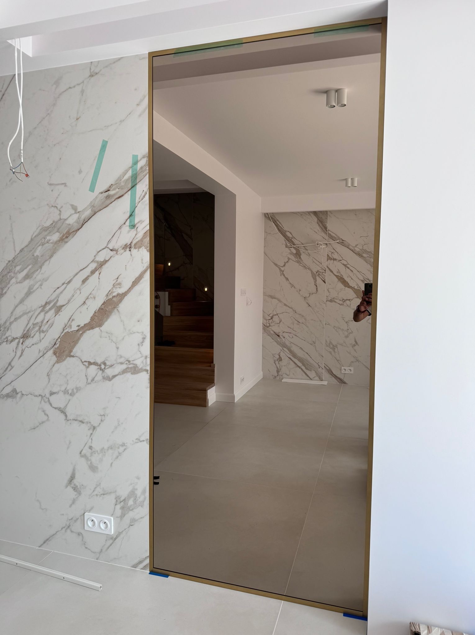 Szklana ściana działowa z złotą ramą oddzielająca pomieszczenia w nowoczesnym wnętrzu, widoczne marmurowe wykończenie ścian i drewniane schody w tle.