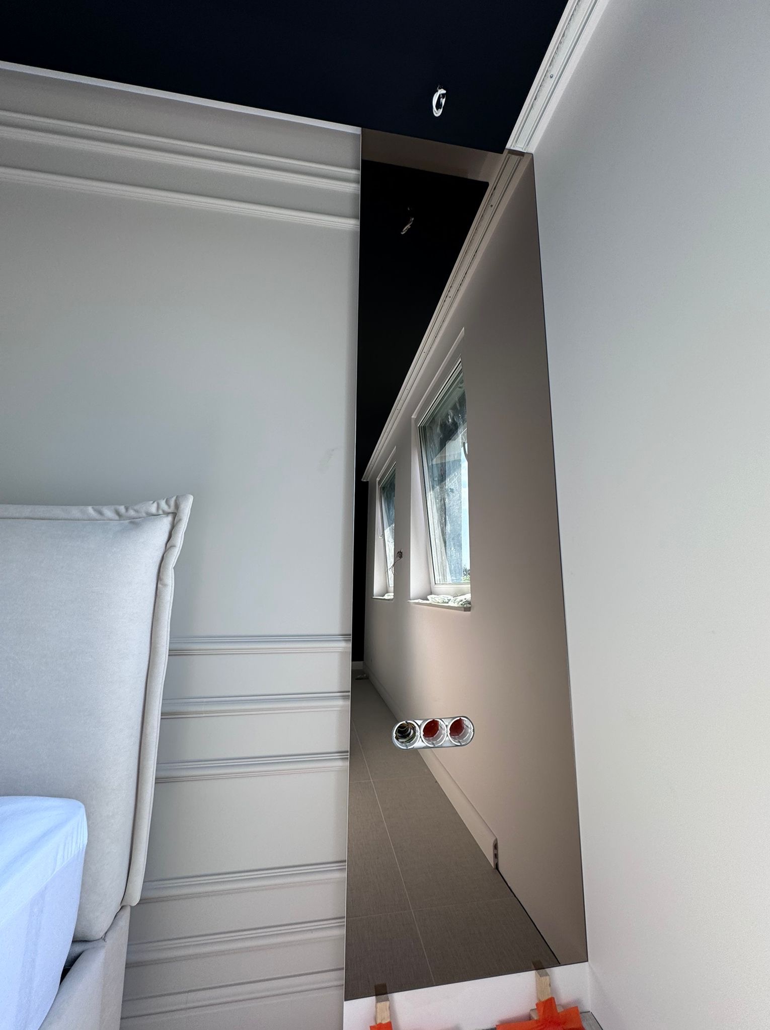 Wnętrze sypialni w trakcie remontu: widoczne okna, fragment łóżka z zagłówkiem, ściany z dekoracyjnymi listwami, instalacja elektryczna i płytki podłogowe w perspektywie.