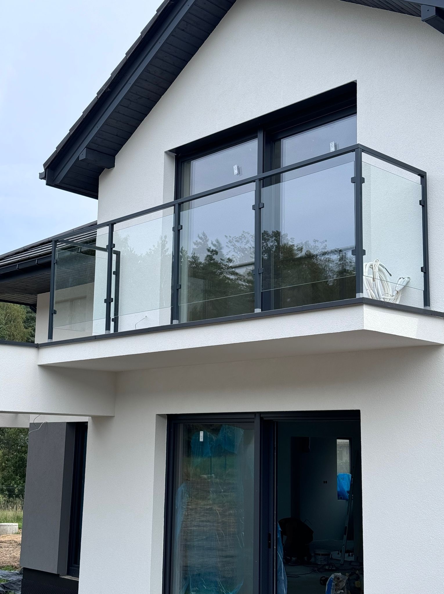 Nowoczesny balkon z czarną ramą i szklaną balustradą w nowym domu z białymi ścianami zewnętrznymi, widoczne przewody na balkonie.