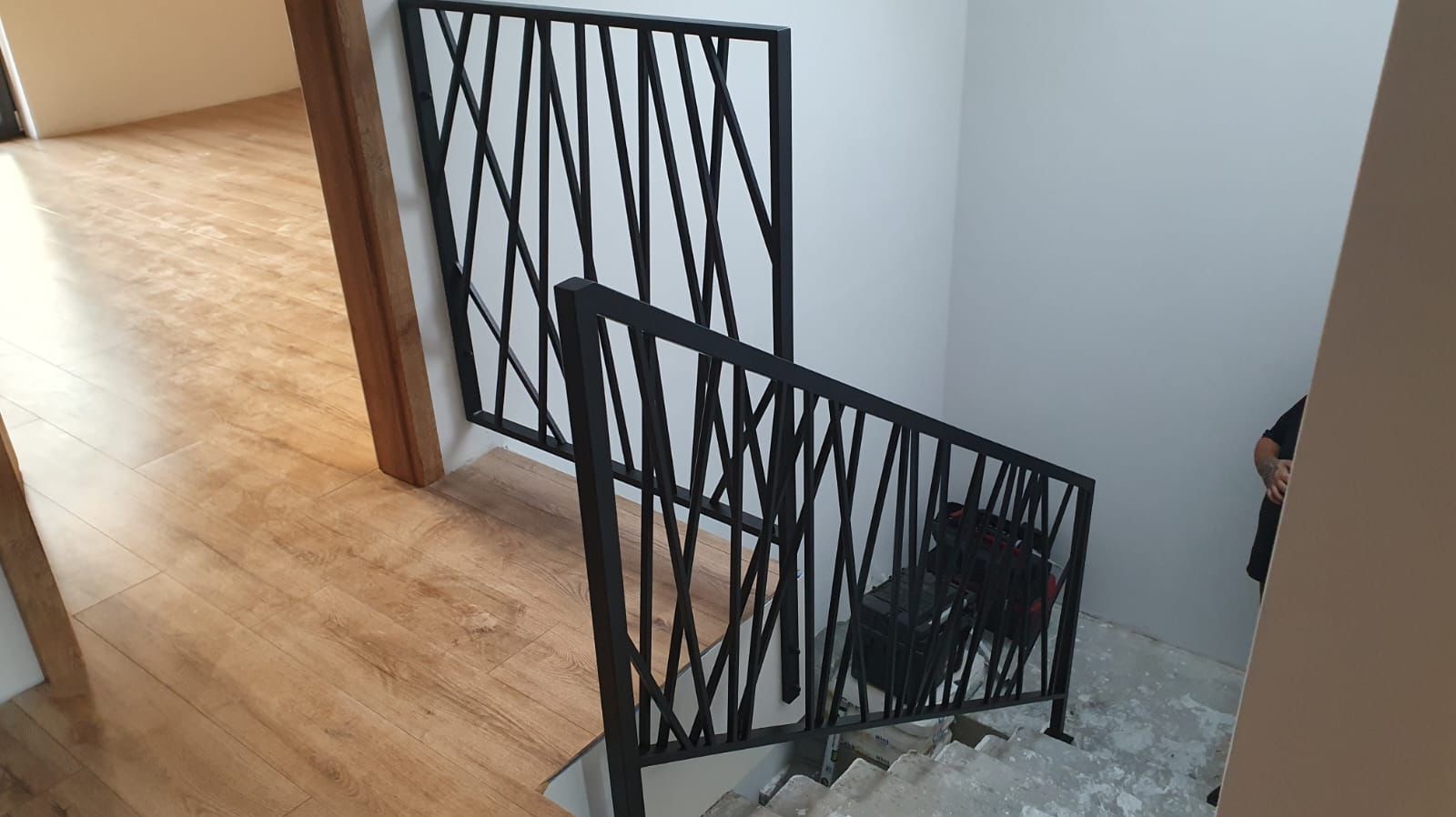 Nowoczesna, czarna balustrada schodowa z geometrycznym wzorem w pionowe, przecinające się linie, widoczna z góry, z podłogą imitującą drewno w tle.