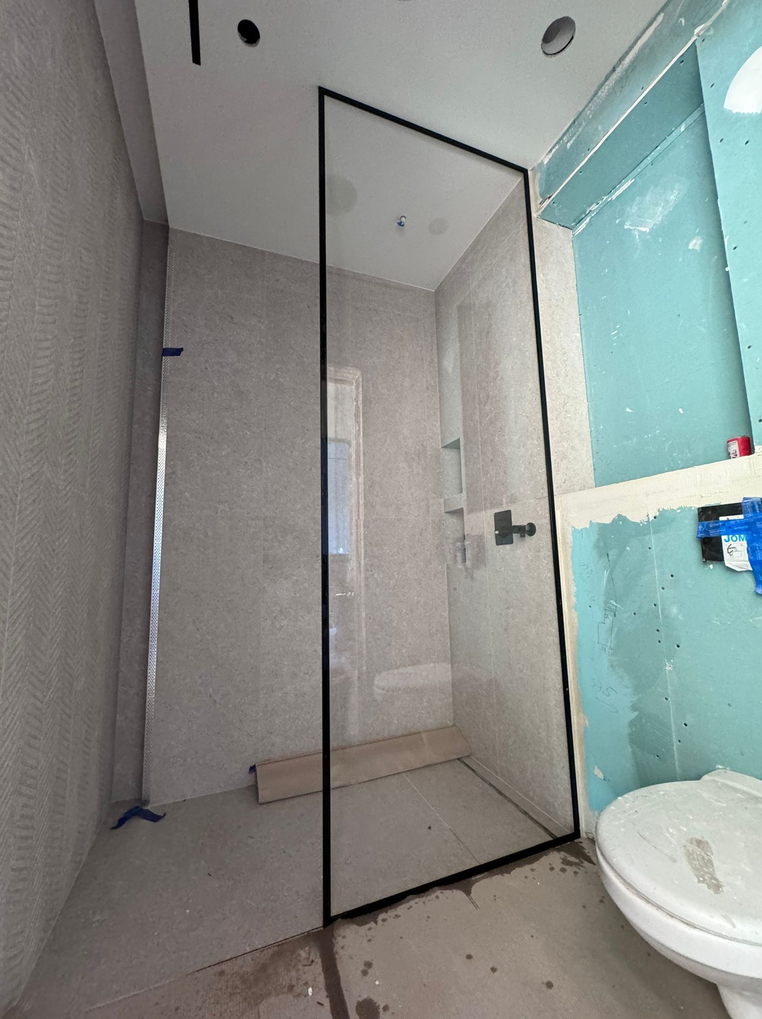 Montaż kabiny prysznicowej z czarną ramą w łazience w trakcie remontu, widoczne płytki, surowa ściana z płyt gipsowo-kartonowych i toaleta.