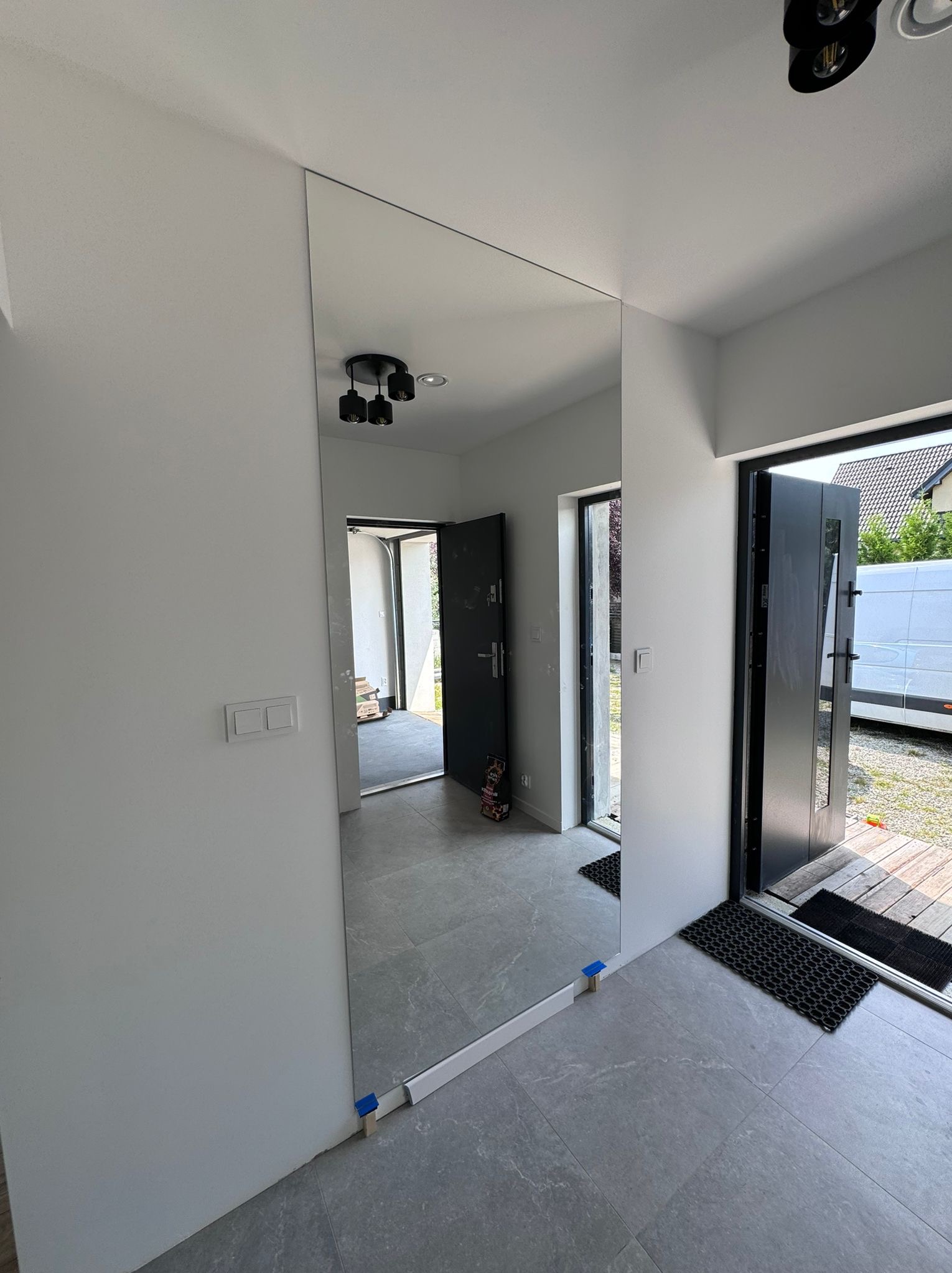 Montaż dużego lustra na ścianie w korytarzu w Sosnowcu, widoczne podpórki i taśma zabezpieczająca podczas instalacji, szare płytki podłogowe i drzwi wejściowe w tle.