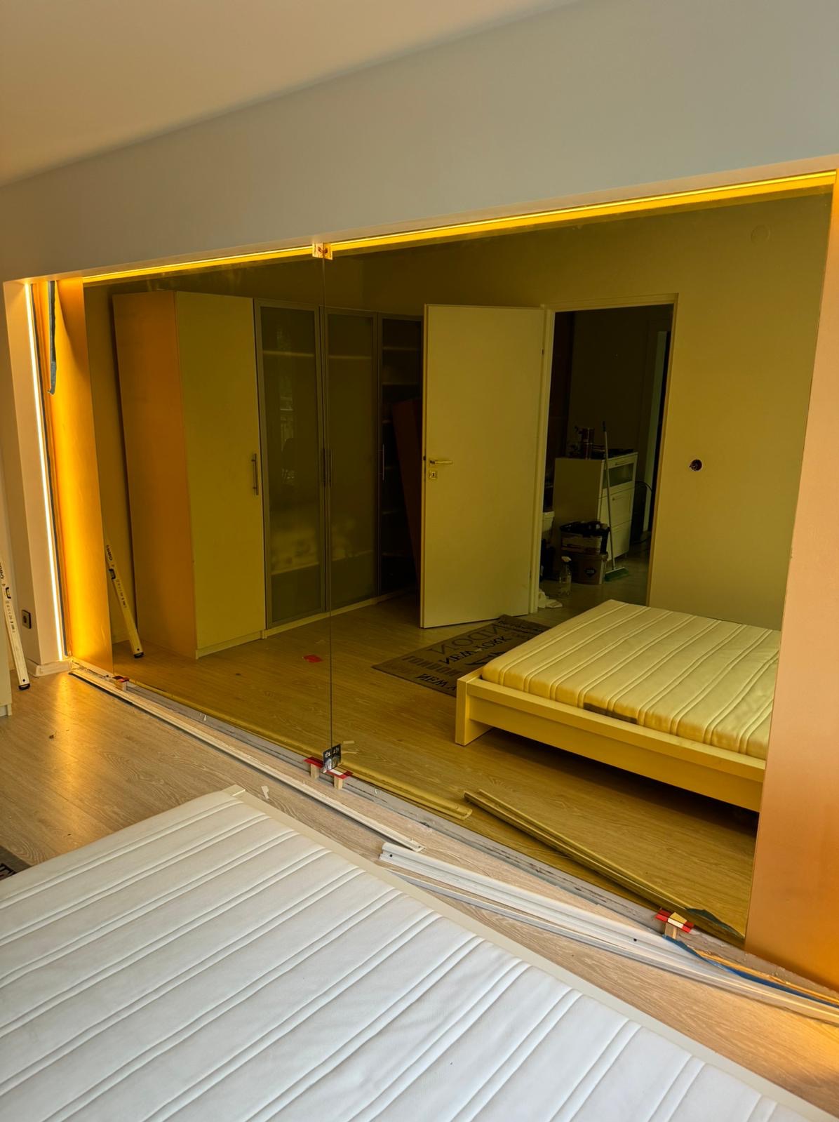 Montaż lustra na ścianie z podświetleniem LED w sypialni, widoczne elementy mocujące i przewody elektryczne.