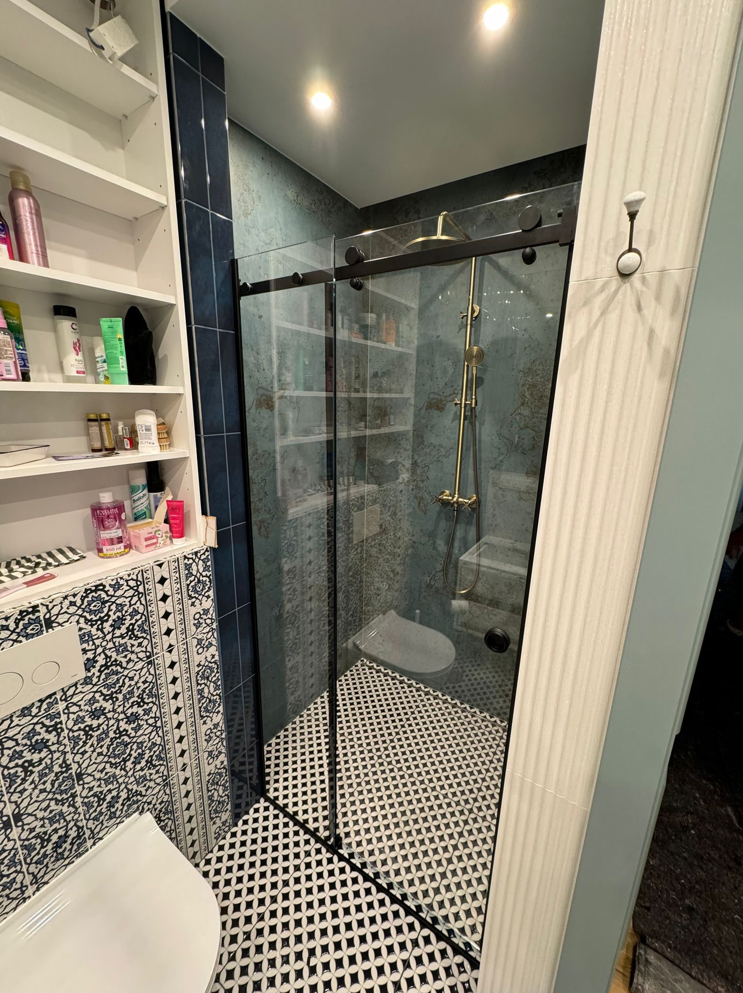 Szklana kabina prysznicowa z przesuwnymi drzwiami, złotą armaturą i czarno-białą mozaiką na podłodze, widok z boku na toaletę i półki z kosmetykami.