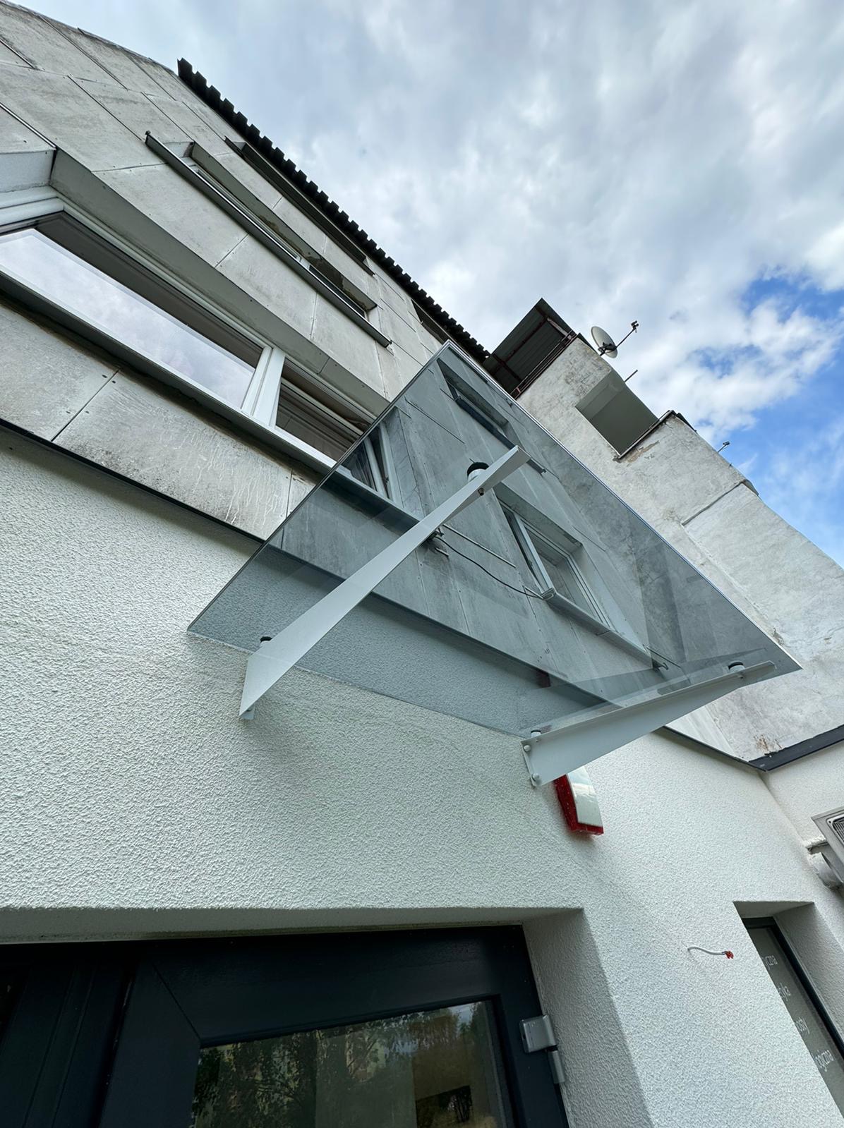 Szklany daszek nad wejściem do budynku z białą elewacją, widok z dołu, niebo z chmurami w tle.