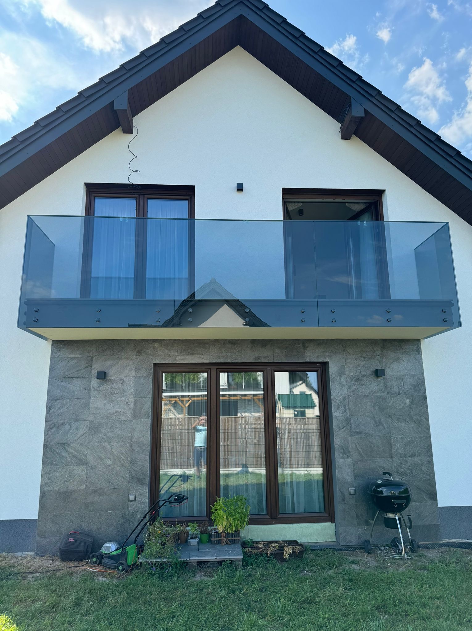 Nowoczesny dom z balkonem zabezpieczonym szklaną balustradą, widoczne mocowania konstrukcji, w tle błękitne niebo.