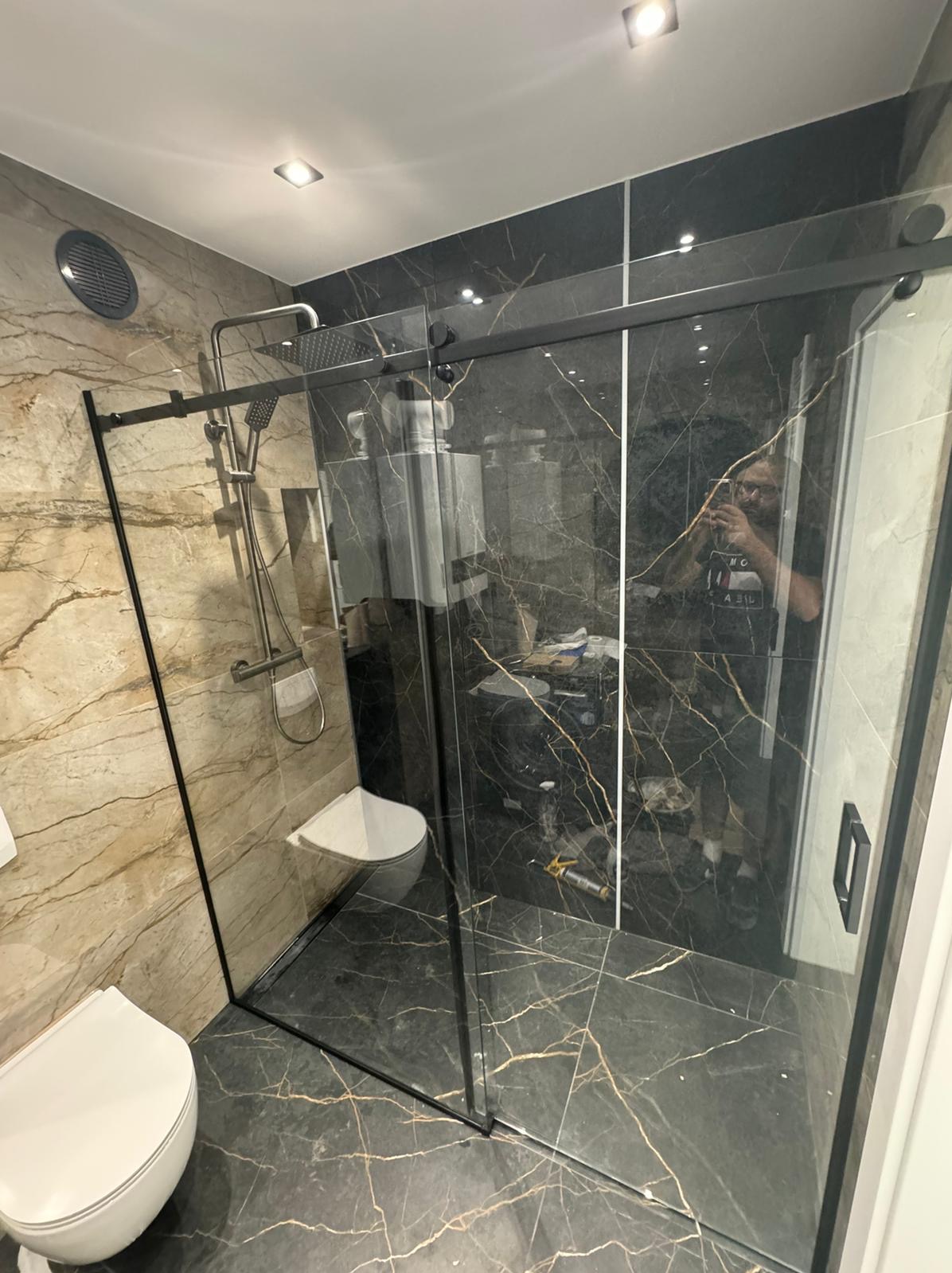 Kabina prysznicowa ze szklanymi drzwiami przesuwnymi w łazience z marmurowymi płytkami, widoczne narzędzia montażowe i odbicie fotografa.