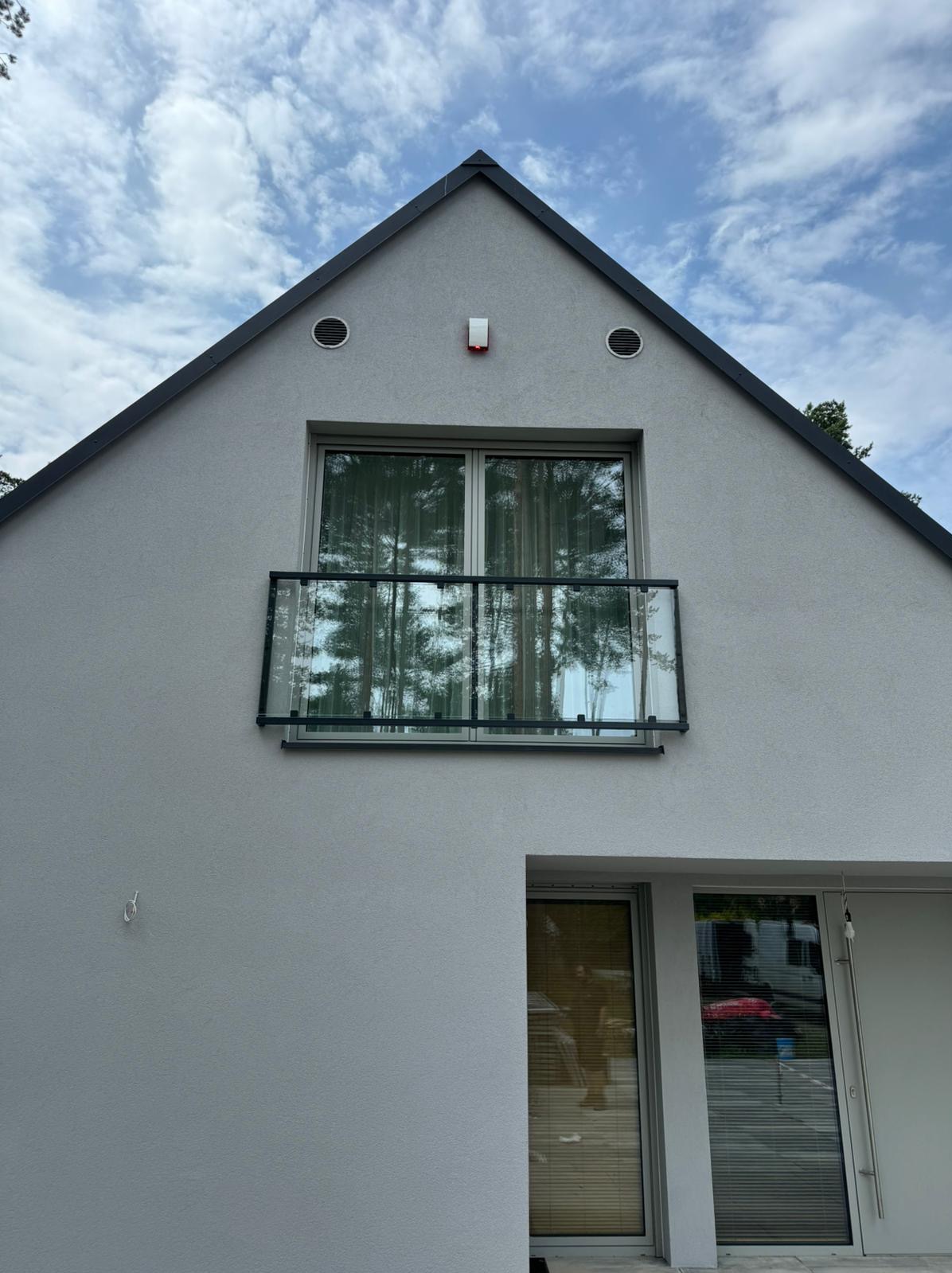 Nowoczesny dom z balkonem z balustradą ze szkła hartowanego, odbijającą drzewa i niebo, widoczne detale architektoniczne jak wentylacja i czujnik alarmowy.