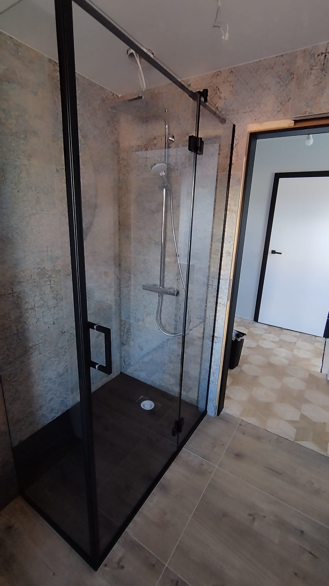 Kabina prysznicowa z czarnymi profilami i szklanymi ścianami, widoczna armatura i odpływ w podłodze, na ścianach i podłodze płytki imitujące kamień i drewno.
